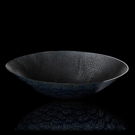 translucent black light grey and dark blue oval bowl perched on a small rounded base with sweeping sides and a fine organic texture moving over the surface resembling ears of corn hand made from glass