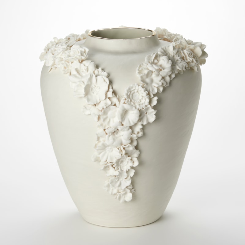 off white cream tall rounded vase with tapering base and narrowing top opening with a band of gold adorned with two lines of three dimensional flowers creating a v formation on the front like the plunging neck line of a ladies dress hand made from porcelain