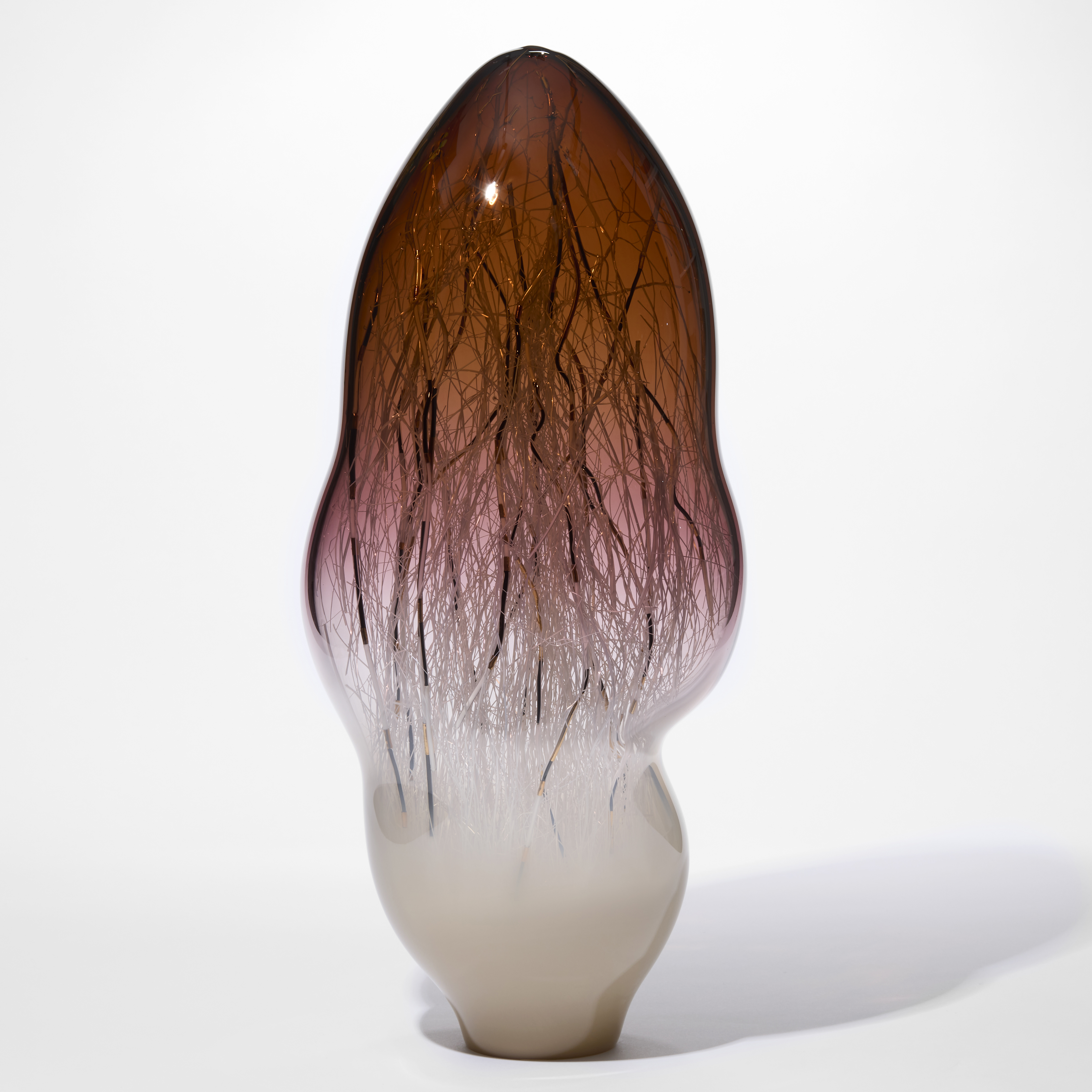 tall undulating amorphic sculpture with the top half in transparent aubergine brown fading towards the base and turning into milky ombre white with the internal filled with long thin canes in white black and gold hand made from blown glass