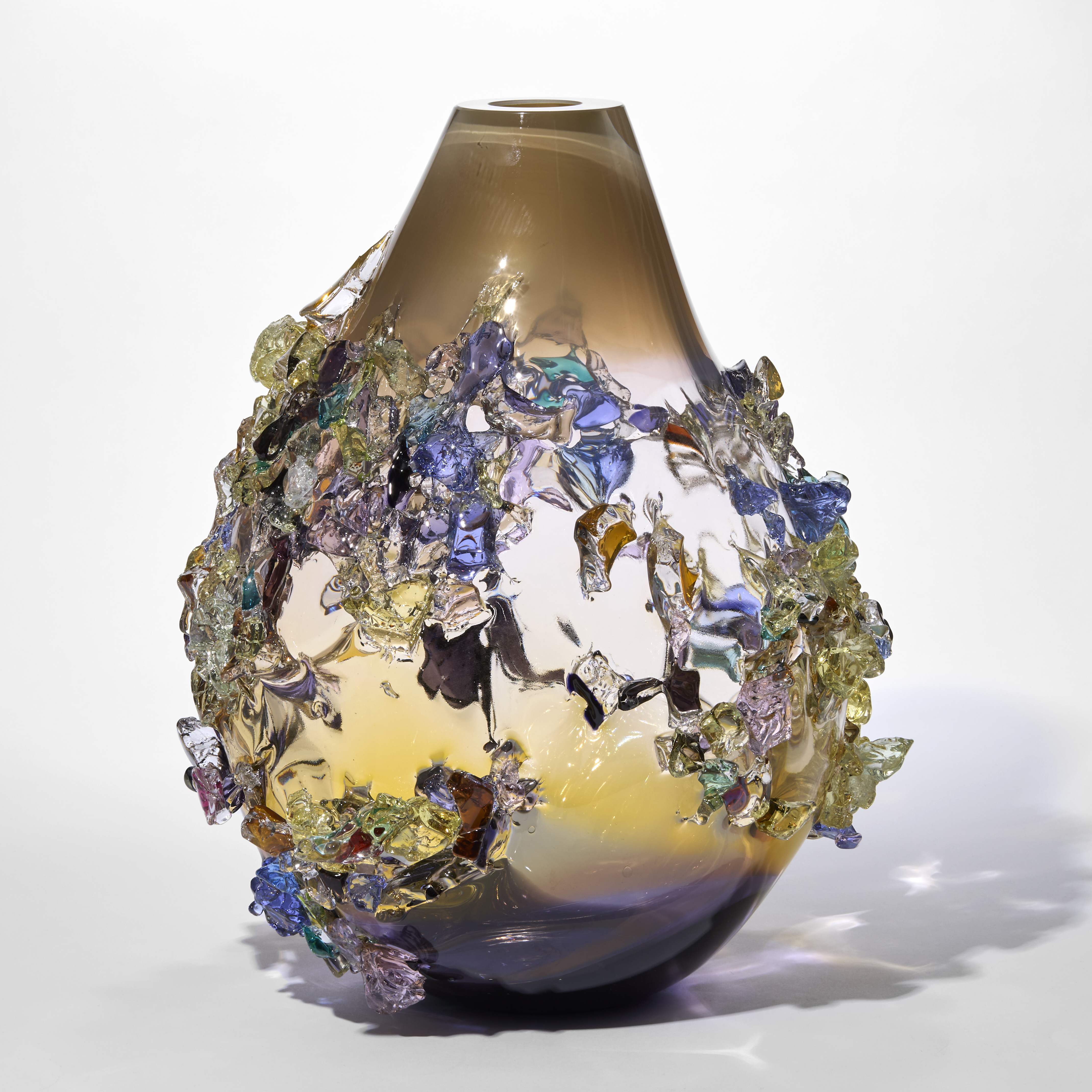 teardrop shaped semi transparent vessel with a dark brown base fading to a soft bronze middle with a milky fawn coloured top neck covered in organic shiny shards in amber yellow pink blue jade and dark brown hand made from glass