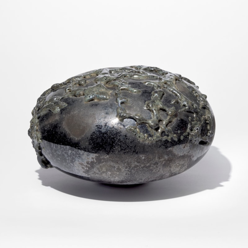 black rounded low mushroom shaped sculpture with leathery weathered surface covered in organic raised surface texture with a dark metallic sheen hand made from glass