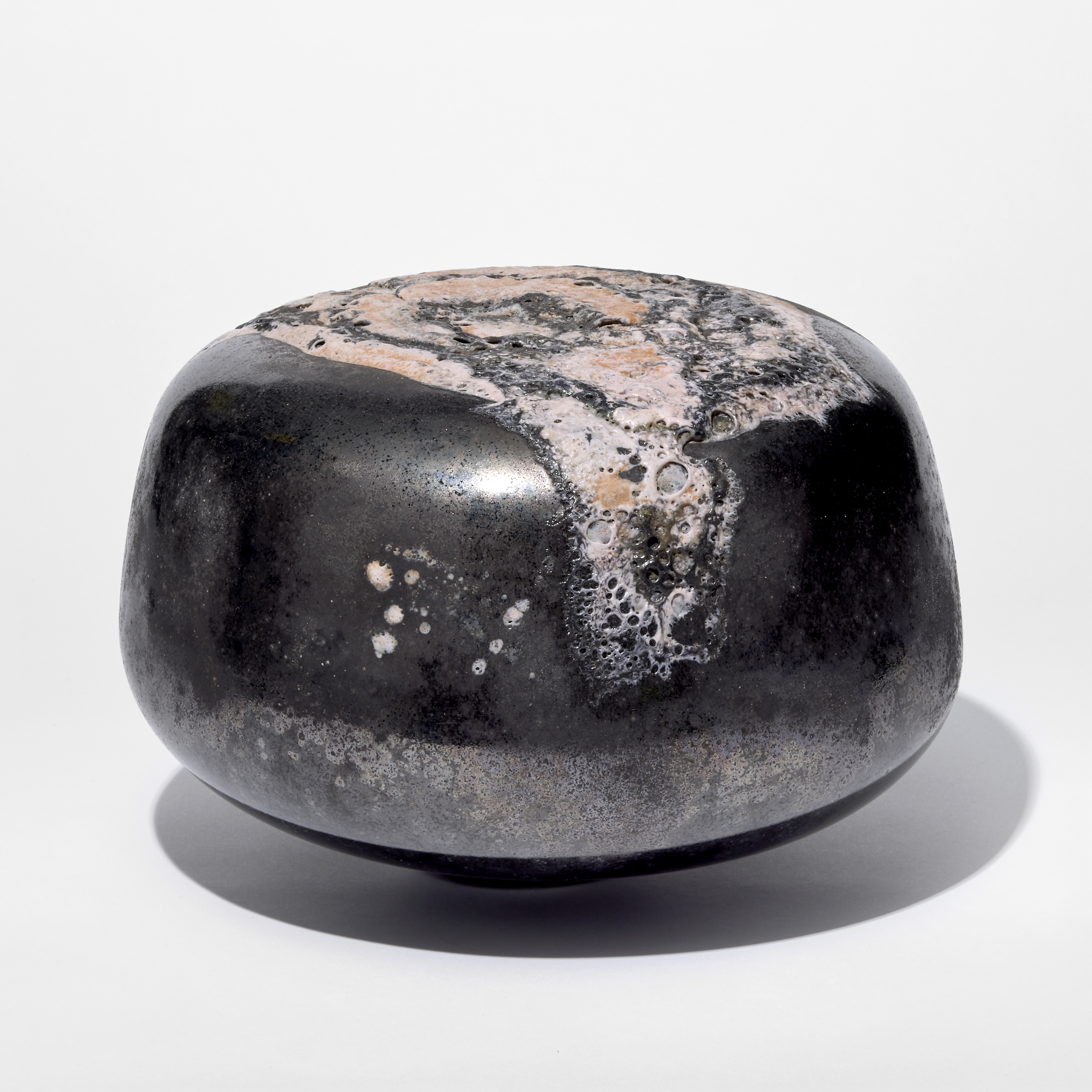 low round solid pebble shaped object with a glossy black pewter finish and raised organic matt pitted areas in soft pink hand made from glass