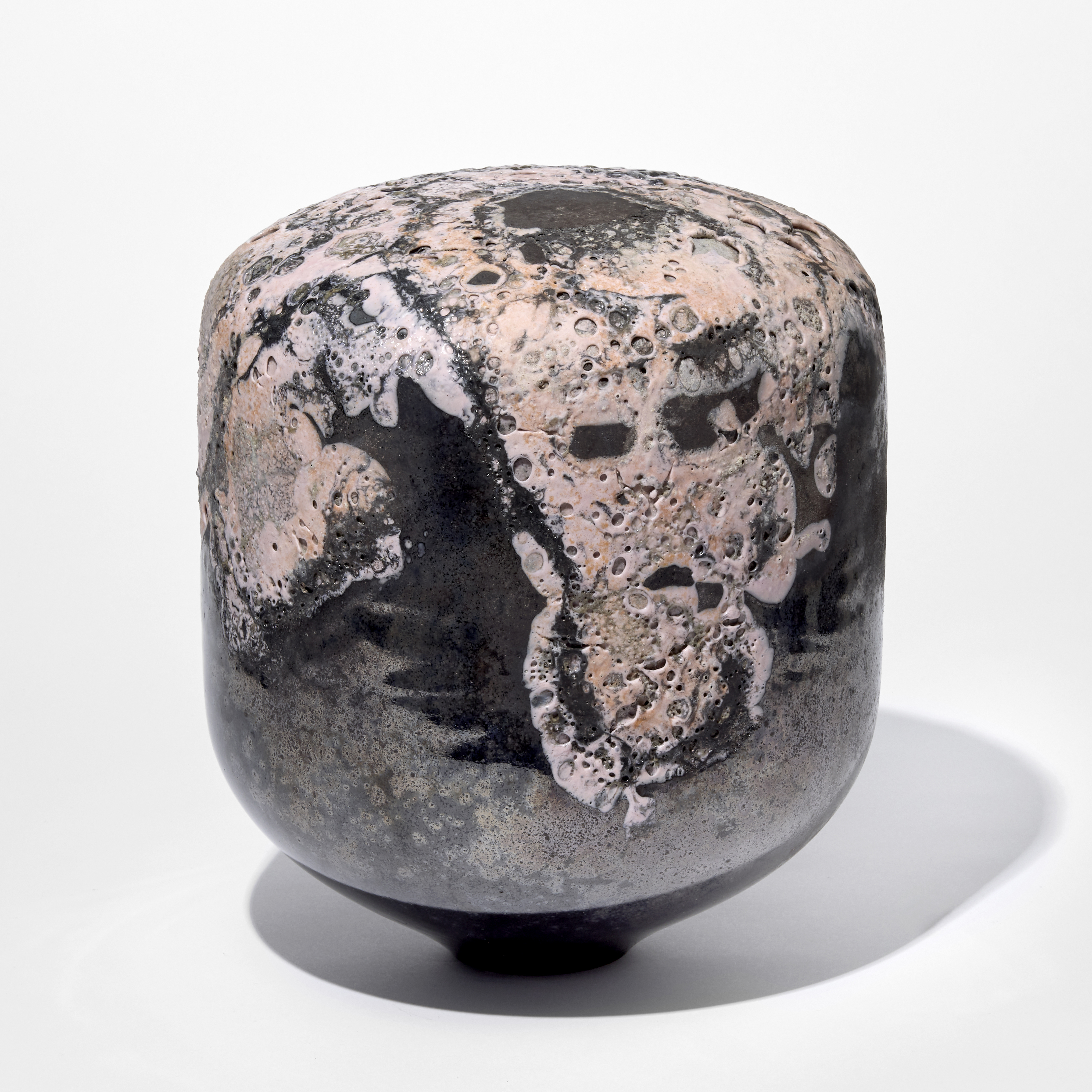 rounded shiny black column shaped solid object with domed top with a raised organic pitted mass on the surface in soft matt pink hand blown from glass 