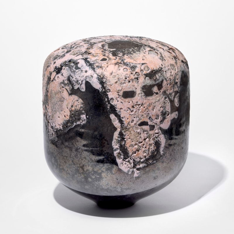 rounded shiny black column shaped solid object with domed top with a raised organic pitted mass on the surface in soft matt pink hand blown from glass
