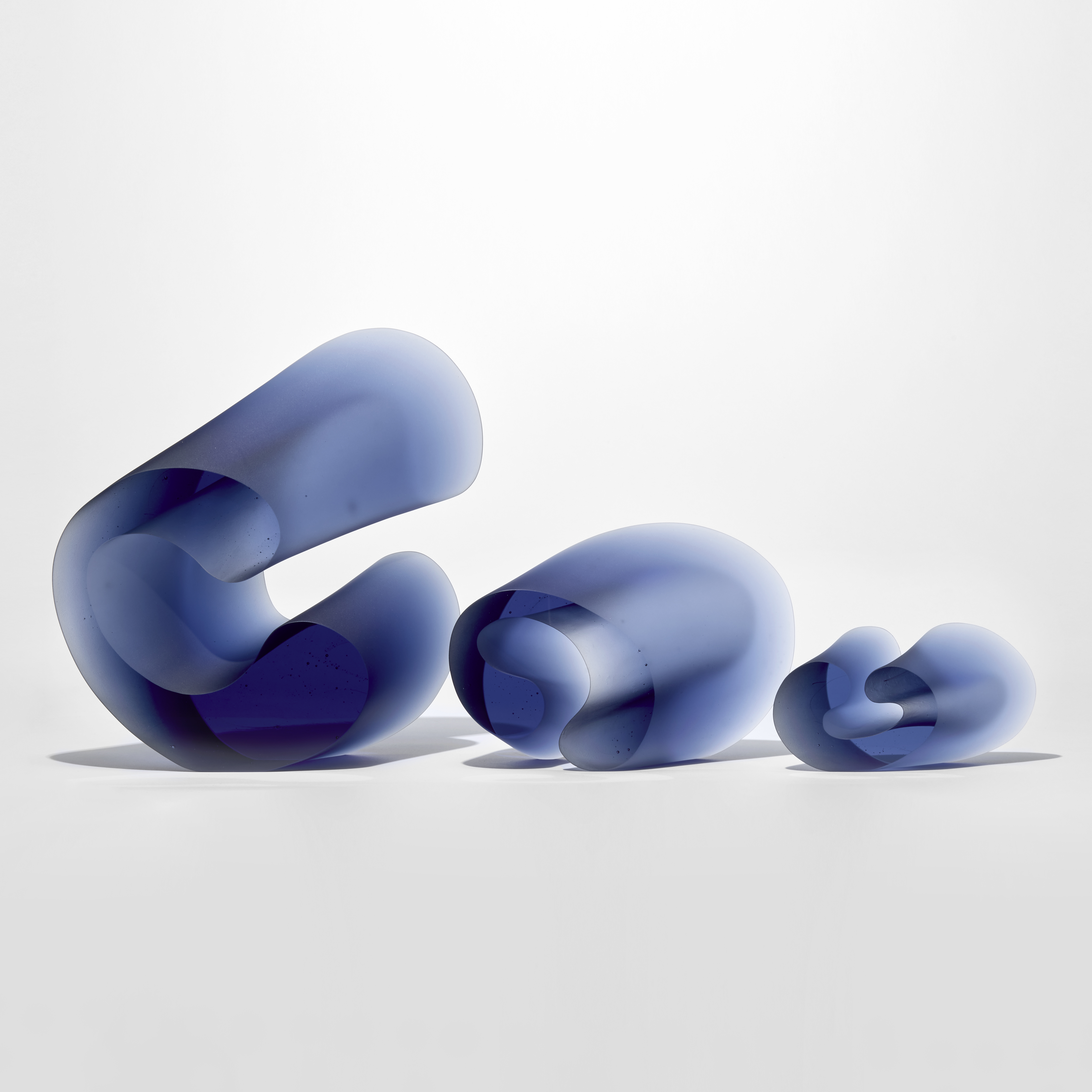 series of three standing curled forms decreasing in size in transparent blue with flat front and back sides with the other sides angled and undulating hand made from glass