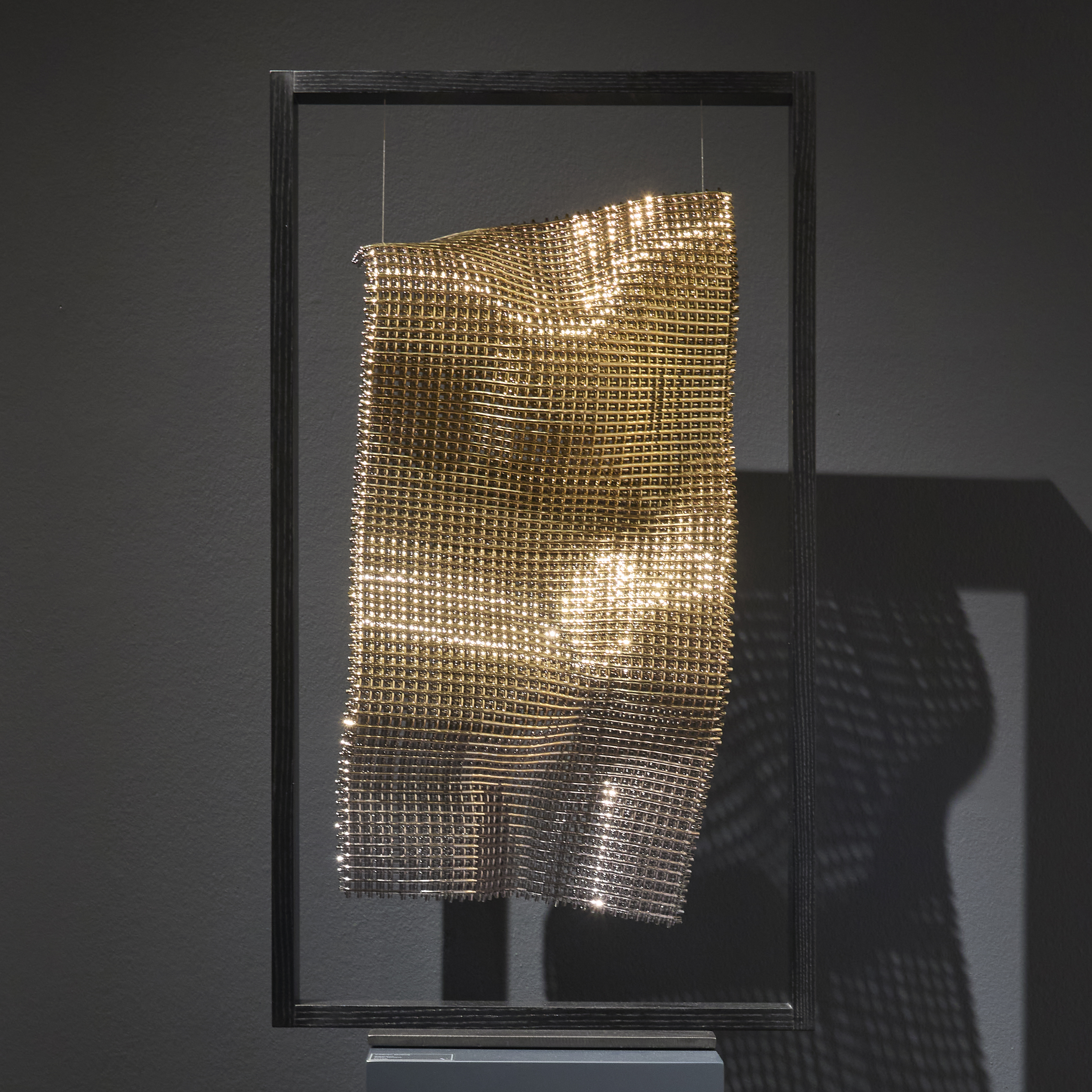 black wooden portrait standing rectangular frame holding a suspended undulating long sheet of cloth made from thick interwoven canes of glass with a gold finish