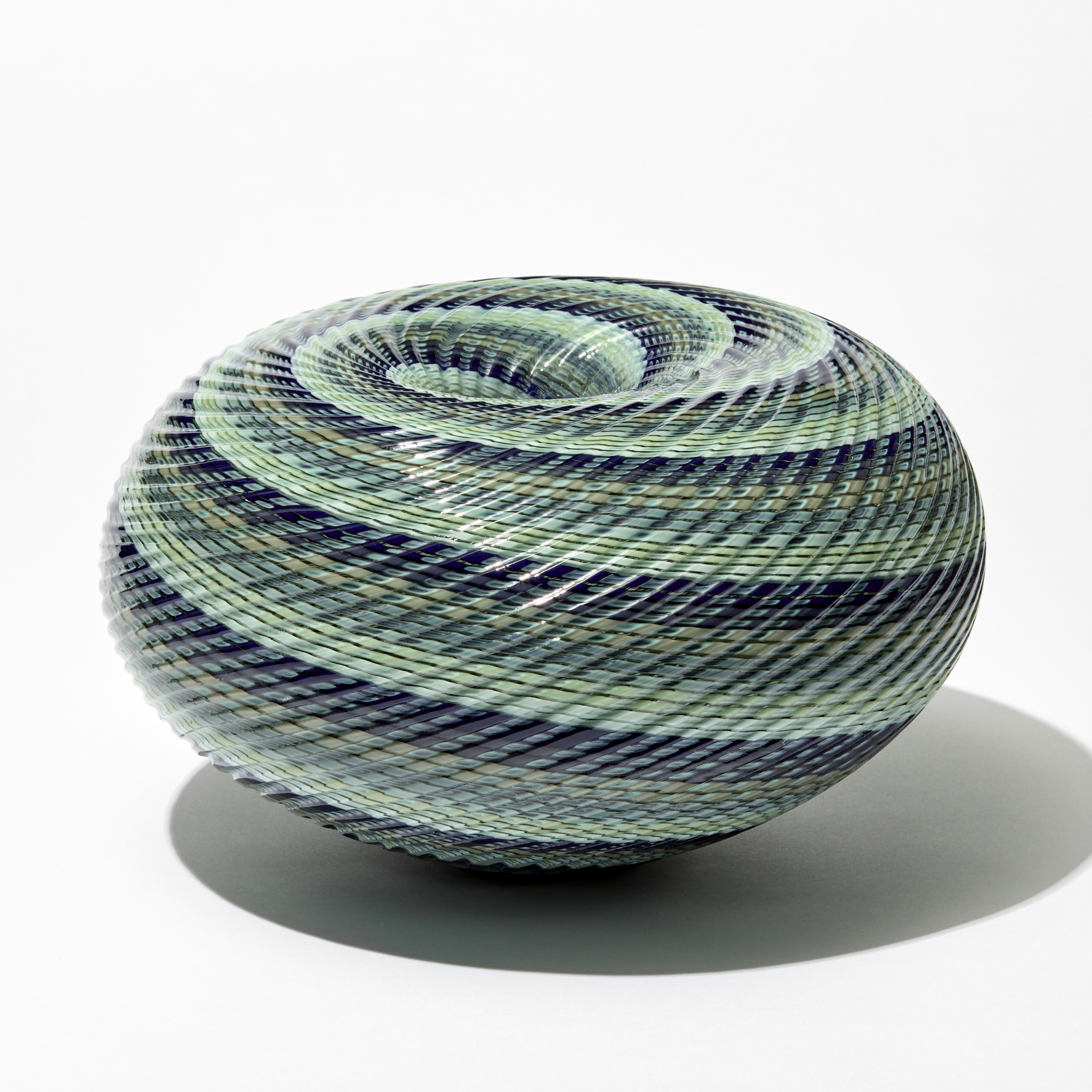 shiny ridged low round maelstrom whirlpool inspired sculpture consisting of thin spiralling bands of jade celadon blue grey and indigo handblown and cut from glass