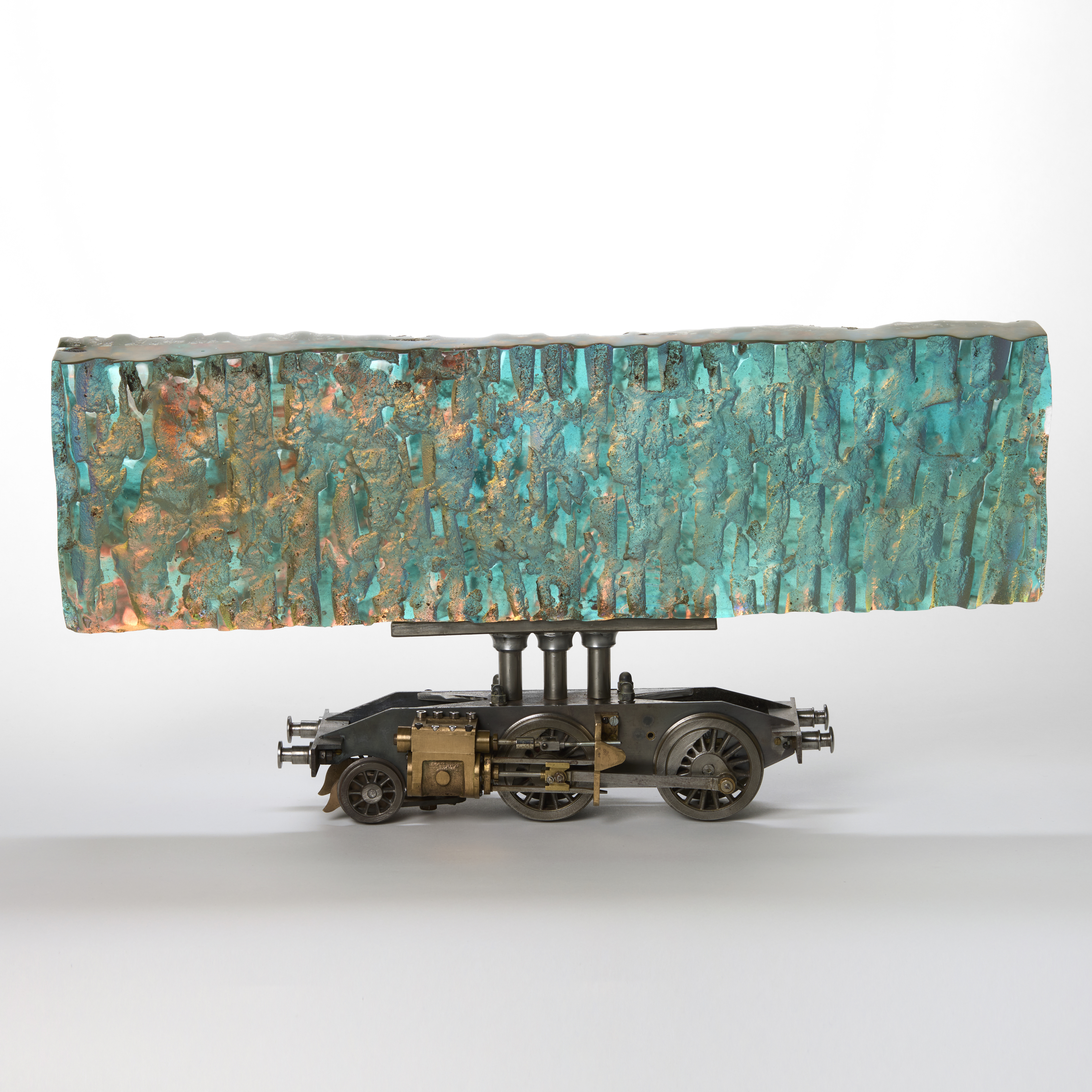 hand made cast glass large thick chunky textured long transparent horizontal block in iridescent colours of pinks peaches turquoises held aloft on a steel model rail way locomotive chassis