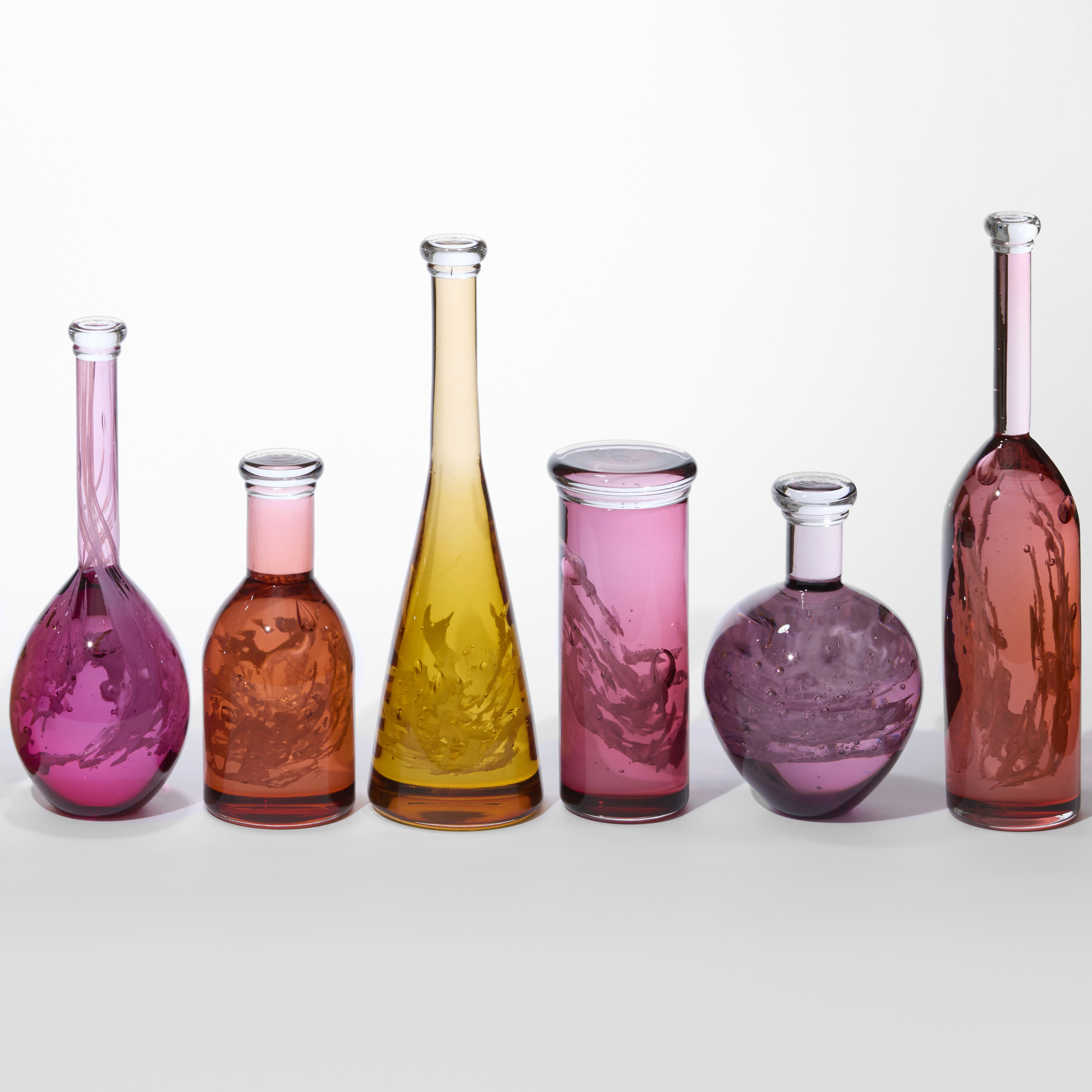 row of ten transparent different shaped bottles with wide bodies and narrow necks with caps on the top with each in red orange yellow peach or coral all containing an intricate lattice coral like structure hand made from glass 