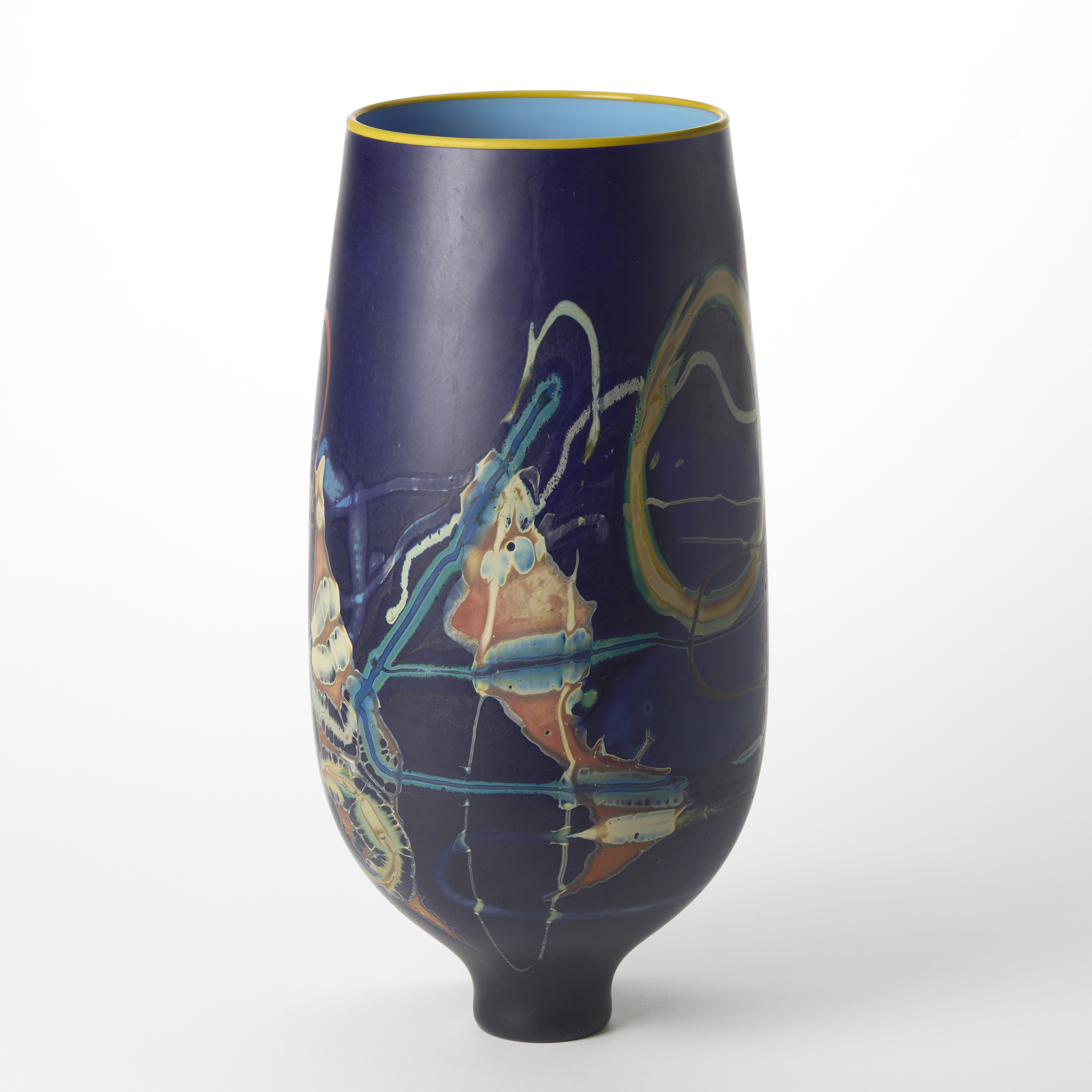 tall rich purple rounded vase perched on a narrow elegant foot with a band of yellow on the top rim and glossy interior in mid blue with the exterior covered in abstract patterns in white pink yellow ochre and teal hand made from glass