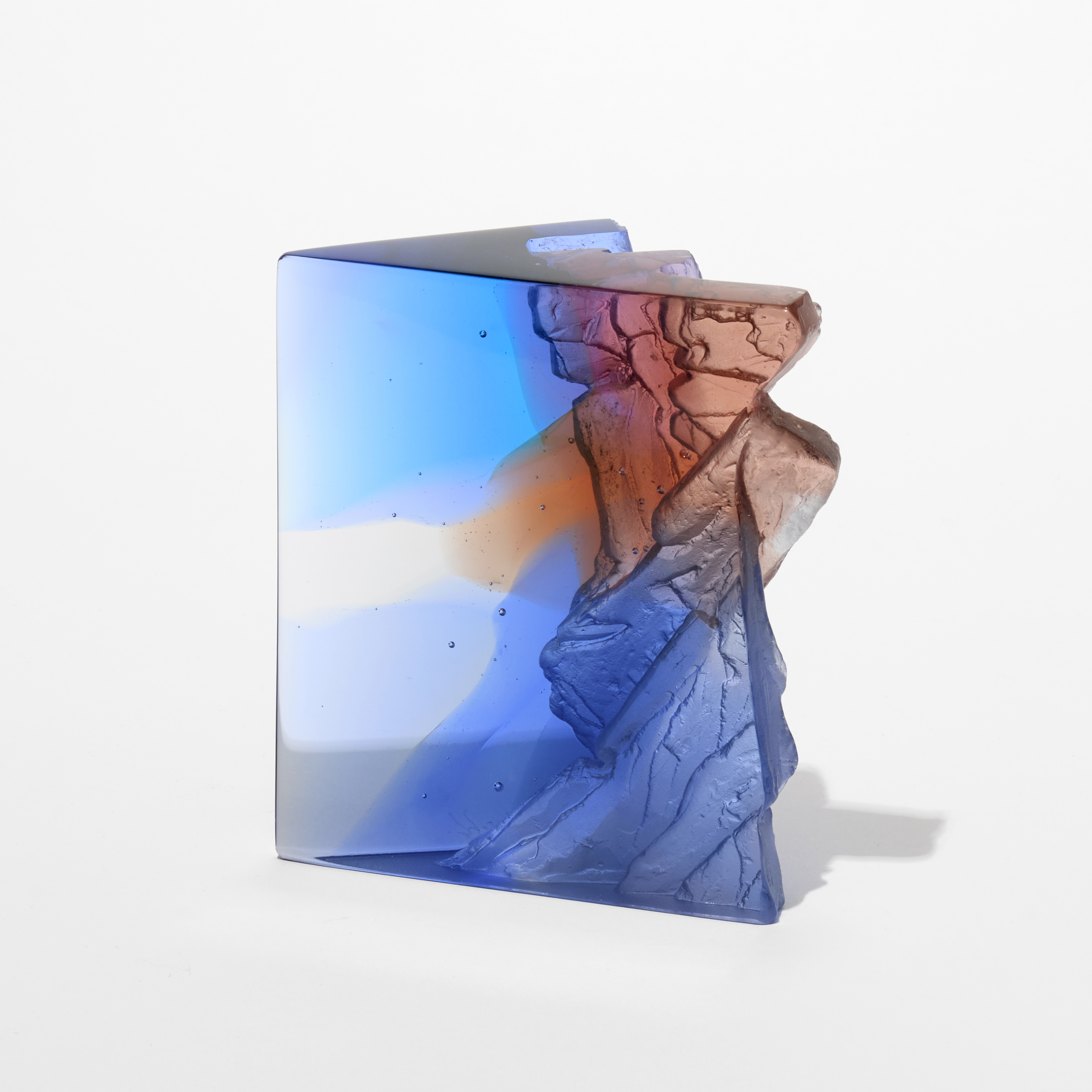transparent blue clear grey and sienna sculpture resembling a section of a cliff face with an angular form and two shiny flat sides with one side covered in craggy texture hand made from glass