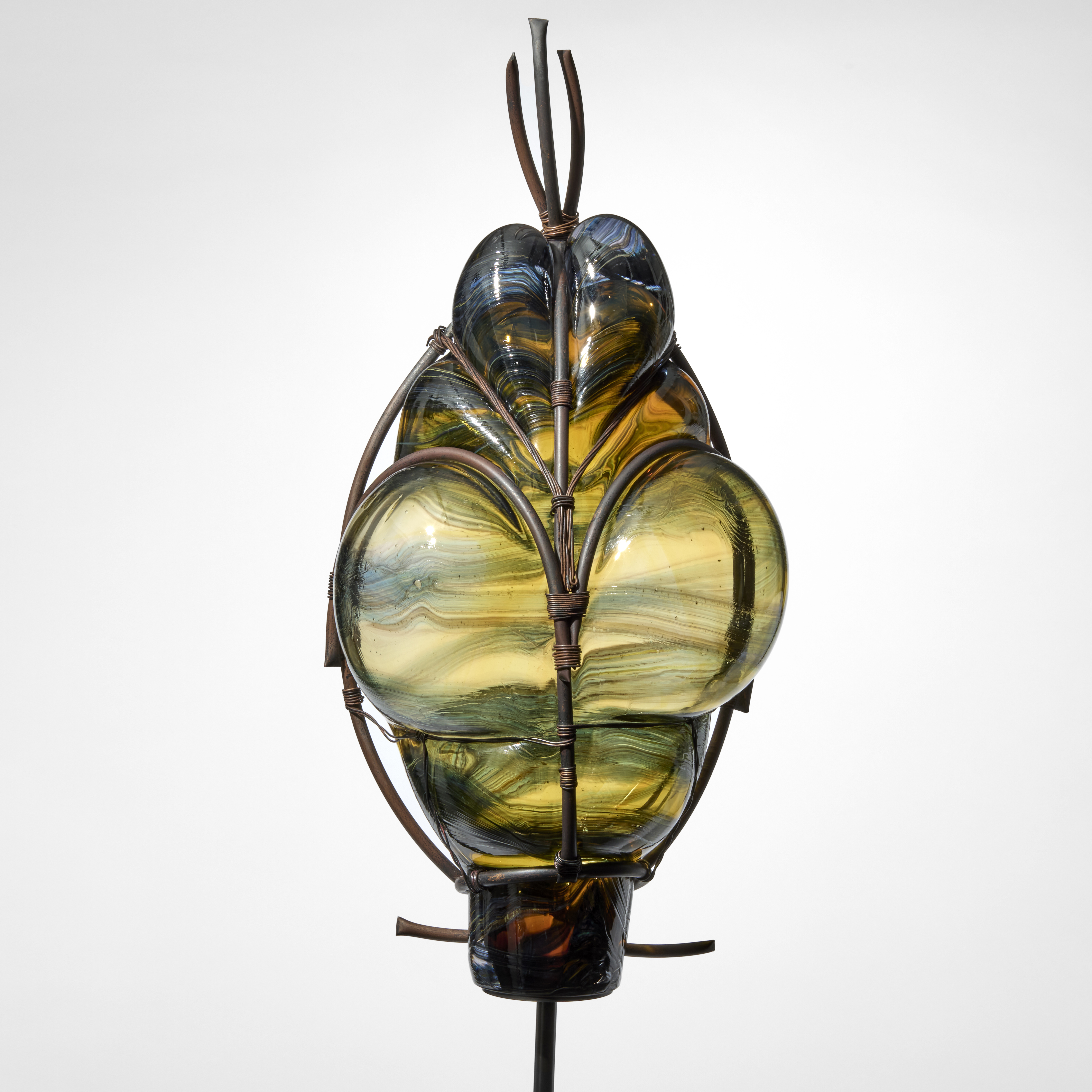 group of standing abstract african tribal masks each raised on a simple blacked steel pole made from merged dark green and blue glass held and bubbling through metal cages created from aged copper pipe