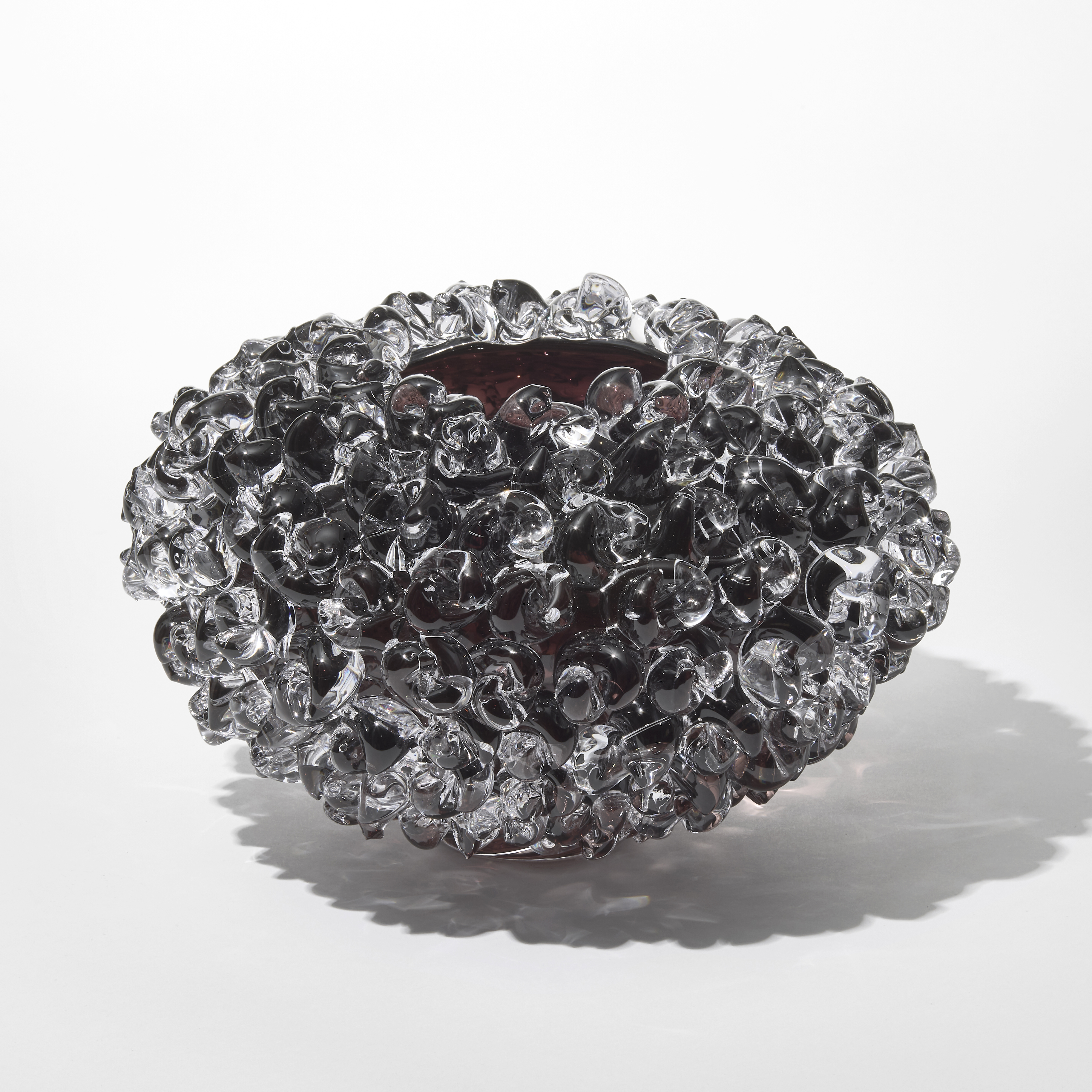 low round discus shaped transparent vase in clear and dark aubergine with the outside covered in raised twisted knotted sections hand made from blown and sculpted glass
