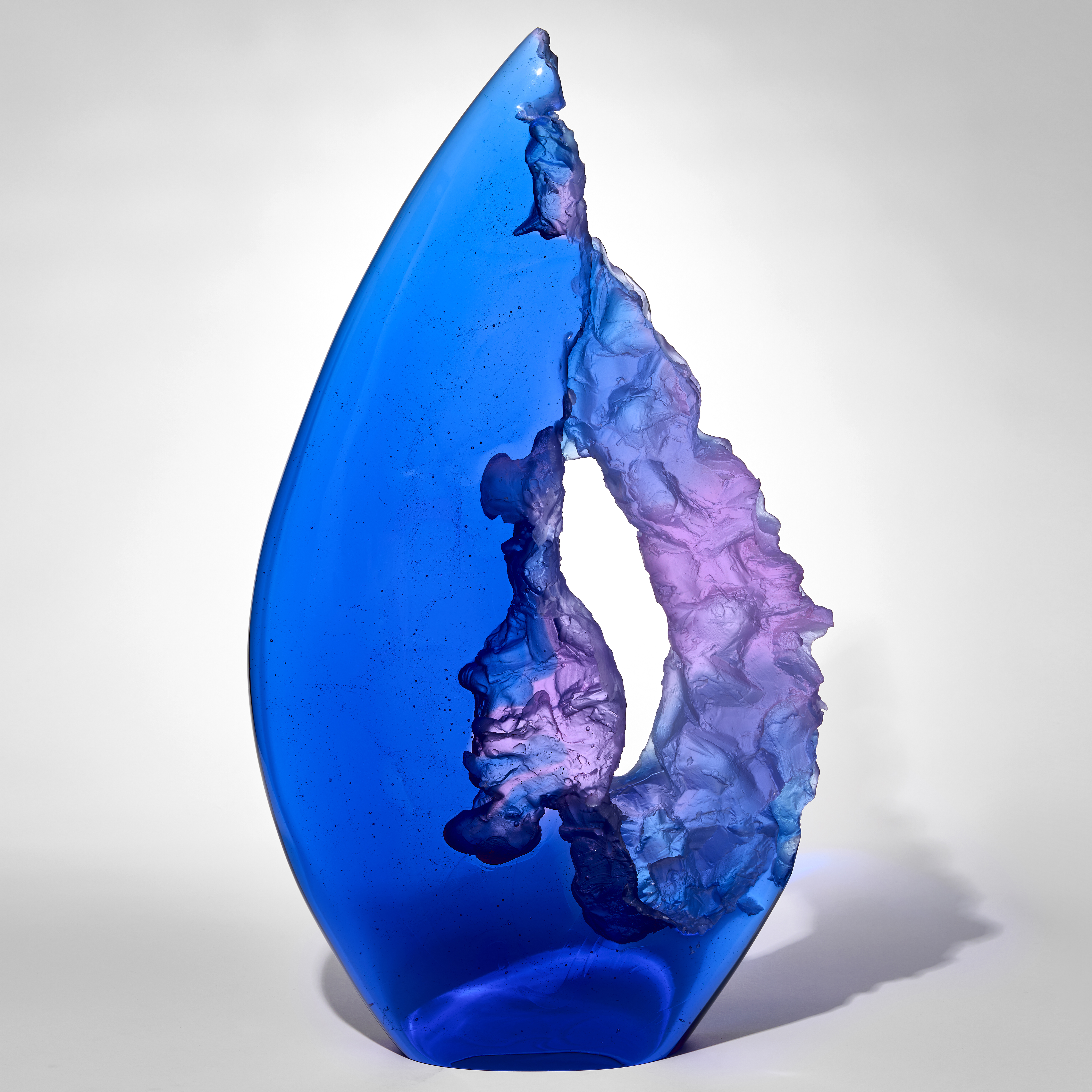 tall pointy transparent teardrop shaped sculpture in electric blue and purple with one side smooth and shiny and the other cut with rock like texture leaving a central opening through the piece hand made from cast glass