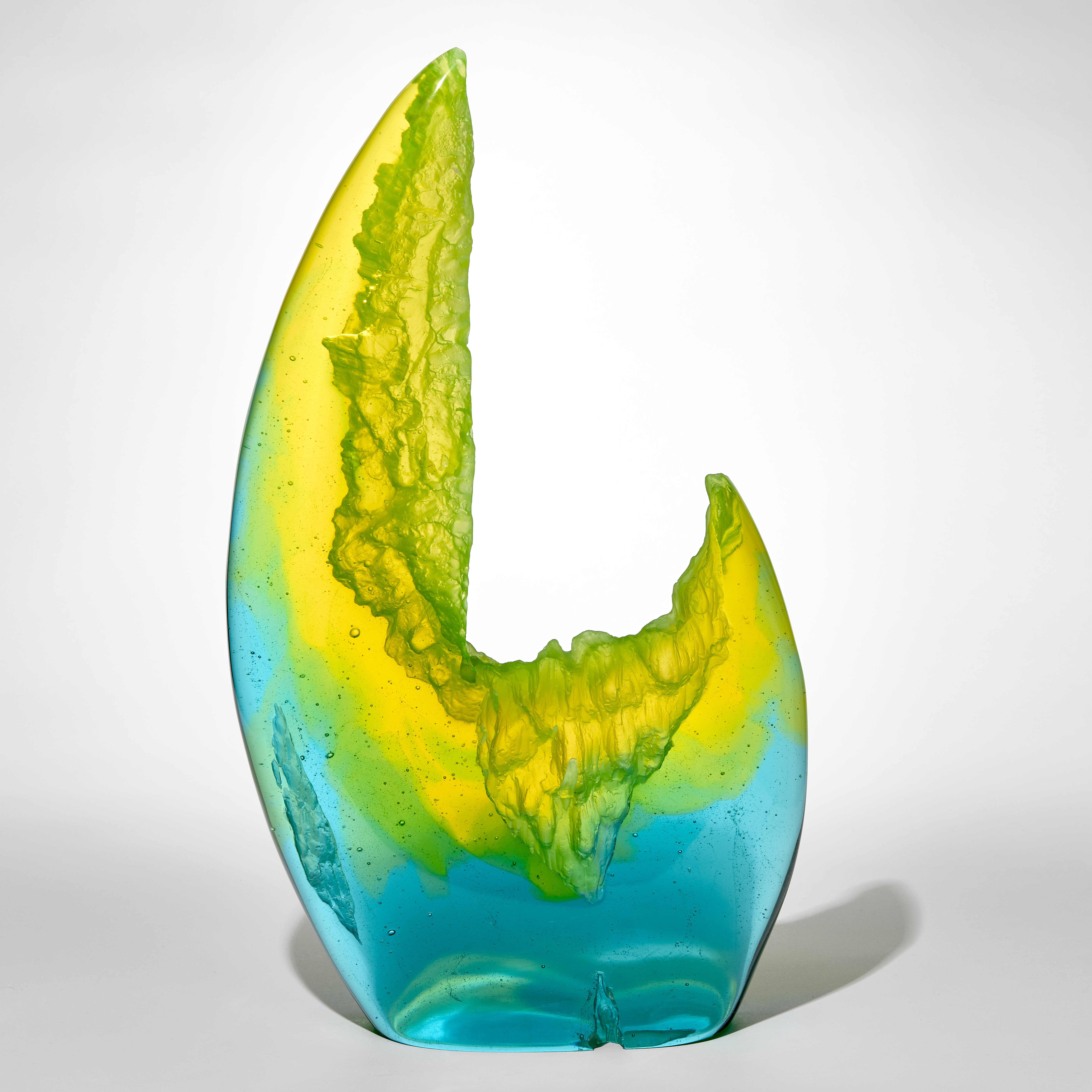 standing transparent rock like form with one side towering higher than the other side in a crab claw like form with bright turquoise at the base merging to lime green and then efflorescent yellow hand made from cast glass