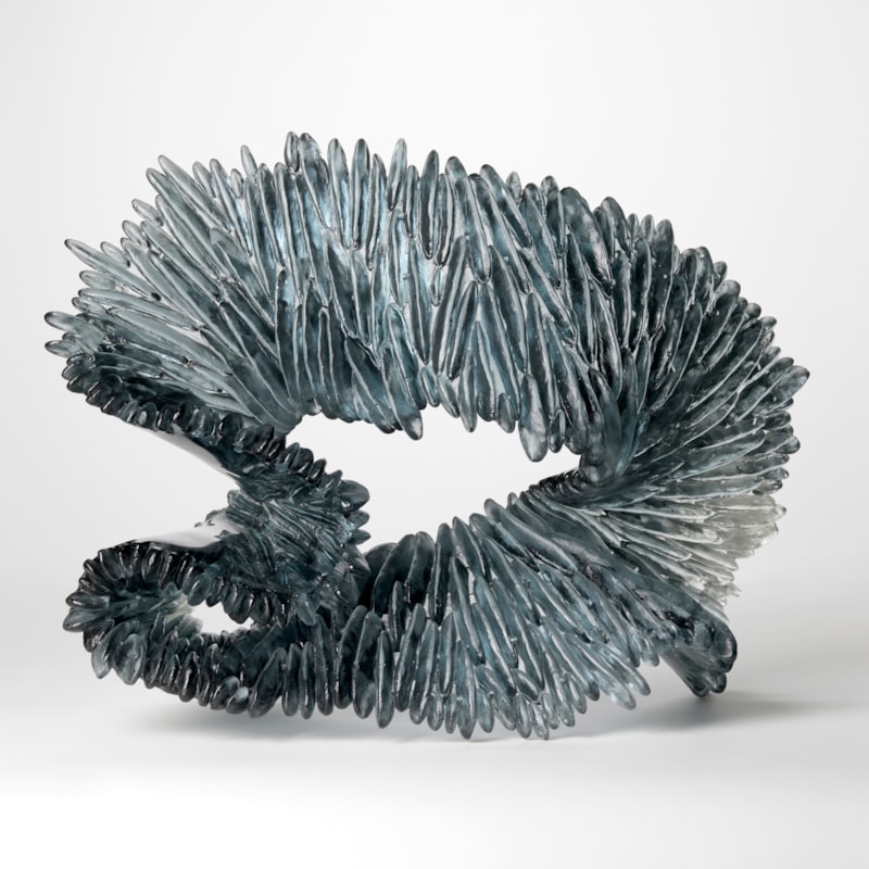 looping standing oval opaque form in rich blue grey and clear with the front surface covered in raised ridges like the gills of a mushroom hand made from cast glass