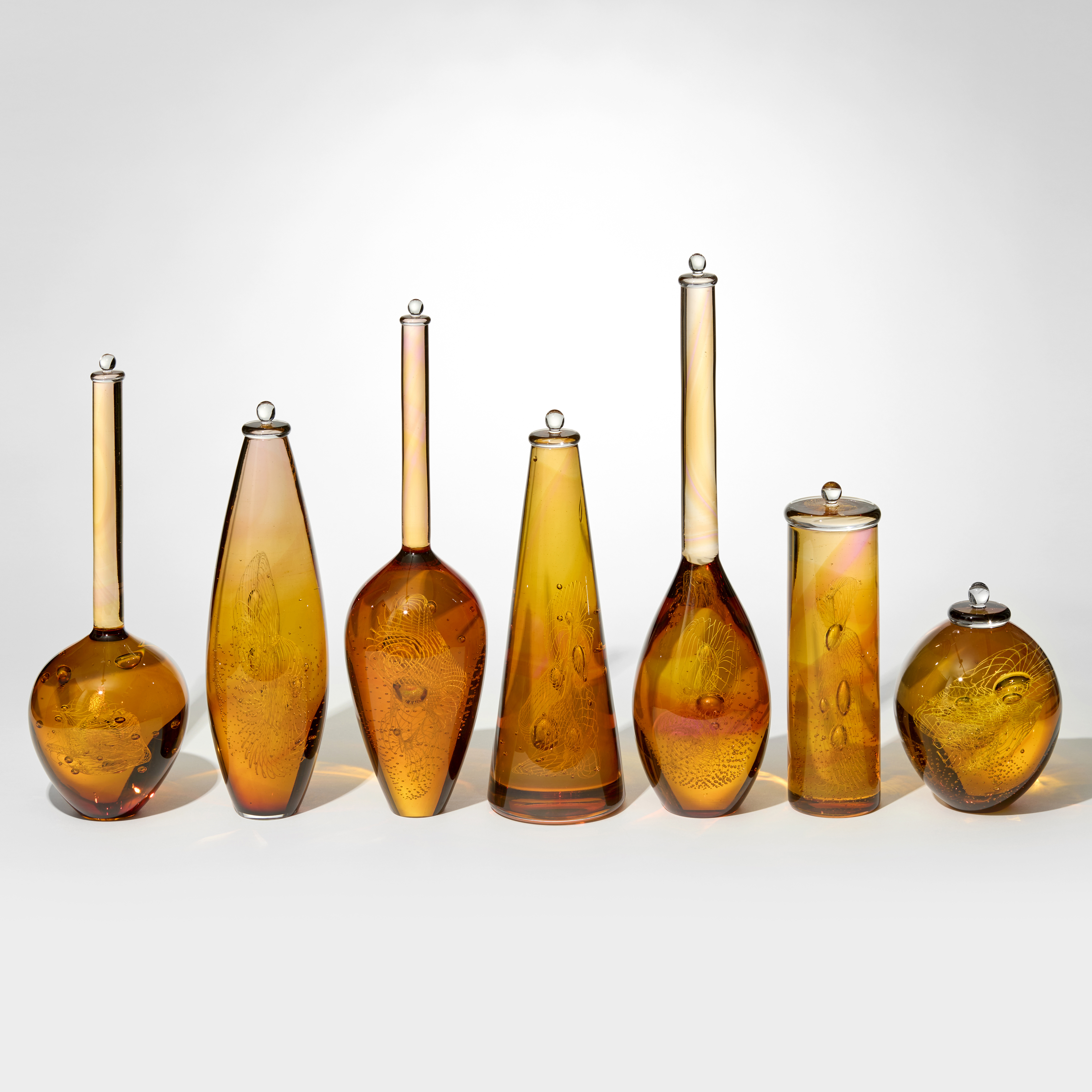 series of seven amber gold and pink transparent bottles in differing shapes with bubbles and patterns trapped within each one hand made from glass