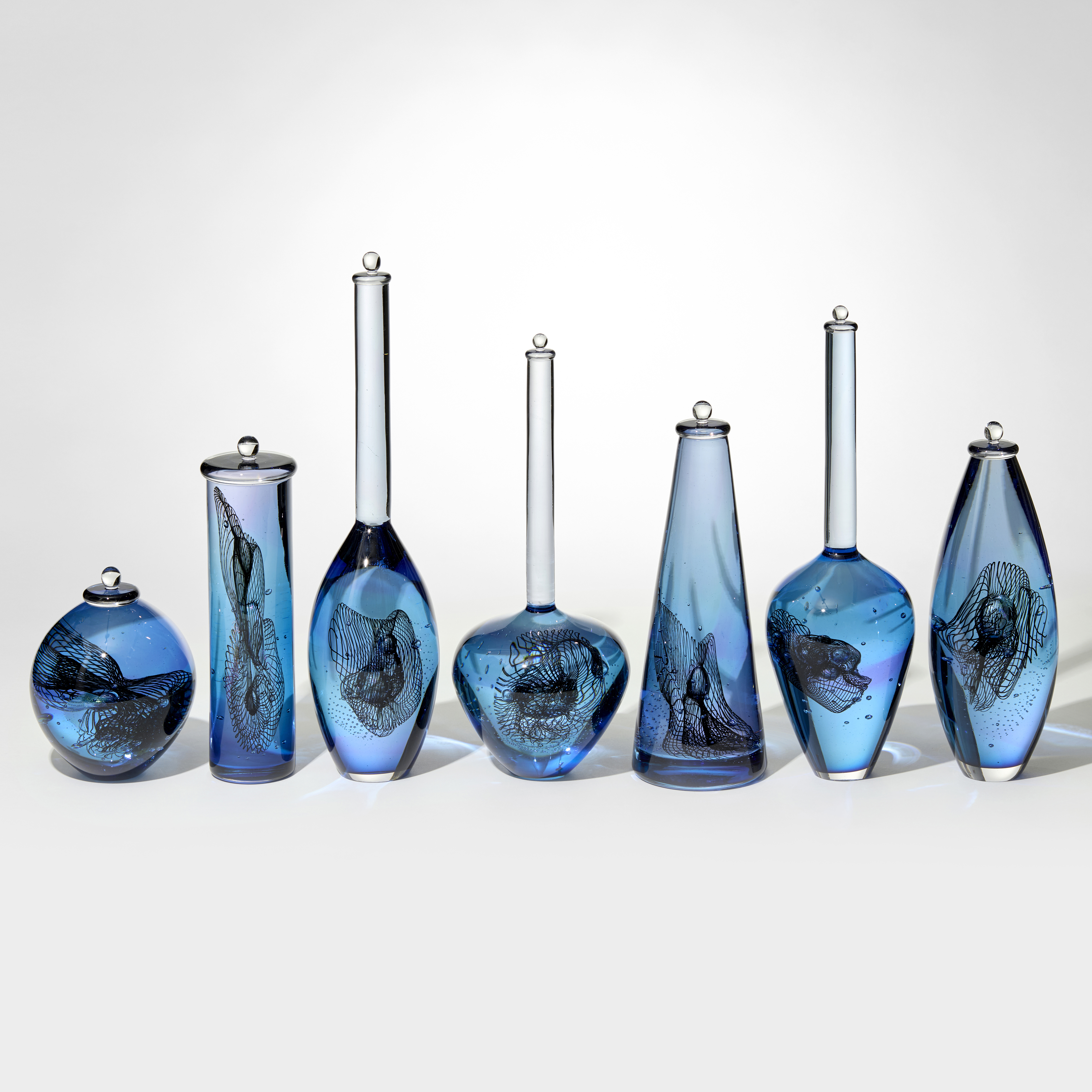 row of seven blue and purple transparent bottles with different shaped bodies and length of long skinny neck with each containing a double helix swirling trapped insert hand blown from glass