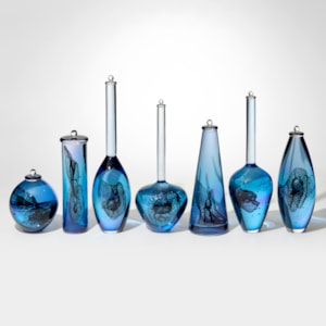row of seven blue and purple transparent bottles with different shaped bodies and length of long skinny neck with each containing a double helix swirling trapped insert hand blown from glass