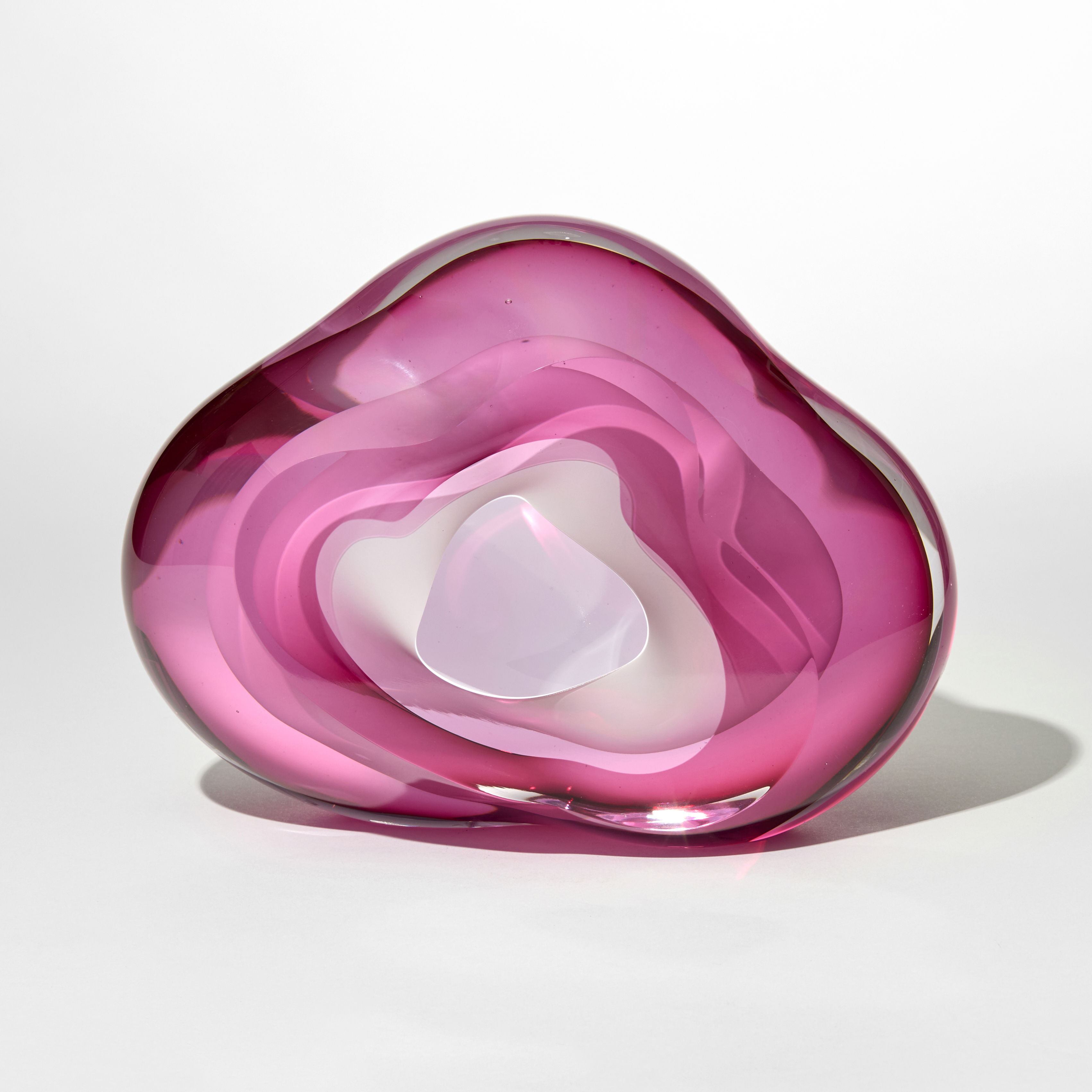 transparent amorphous shaped geode inspired sculpture in layers of clear and bright pink with an opaque white lined inner cavity with the opening on the front hand blown and sculpted from glass