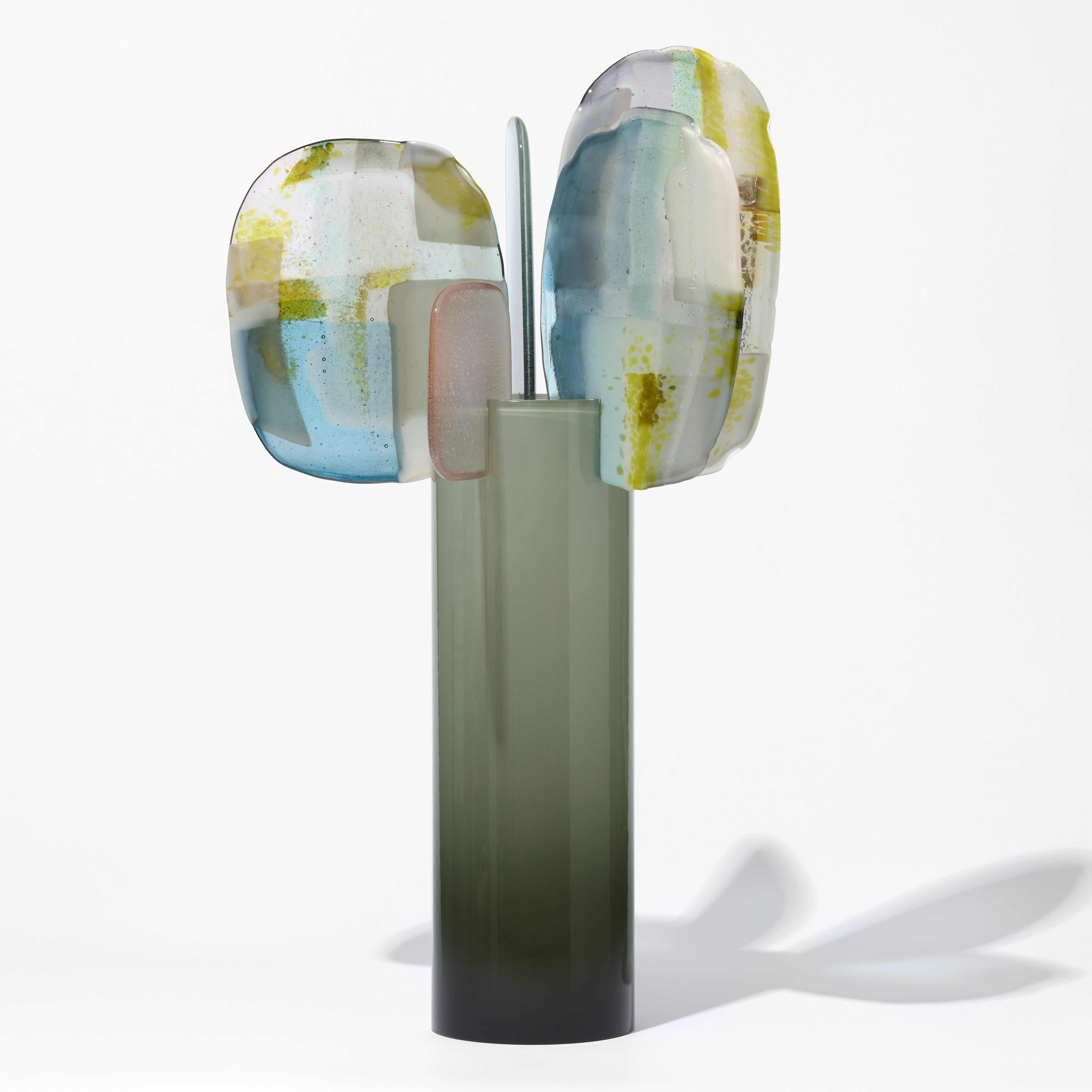 smokey shiny opaque teal cylinder with 5 different shaped and sized rounded petals perched on the top edge each with abstract surface patterns in blue white pink and mustard yellow hand made from blown and fused glass