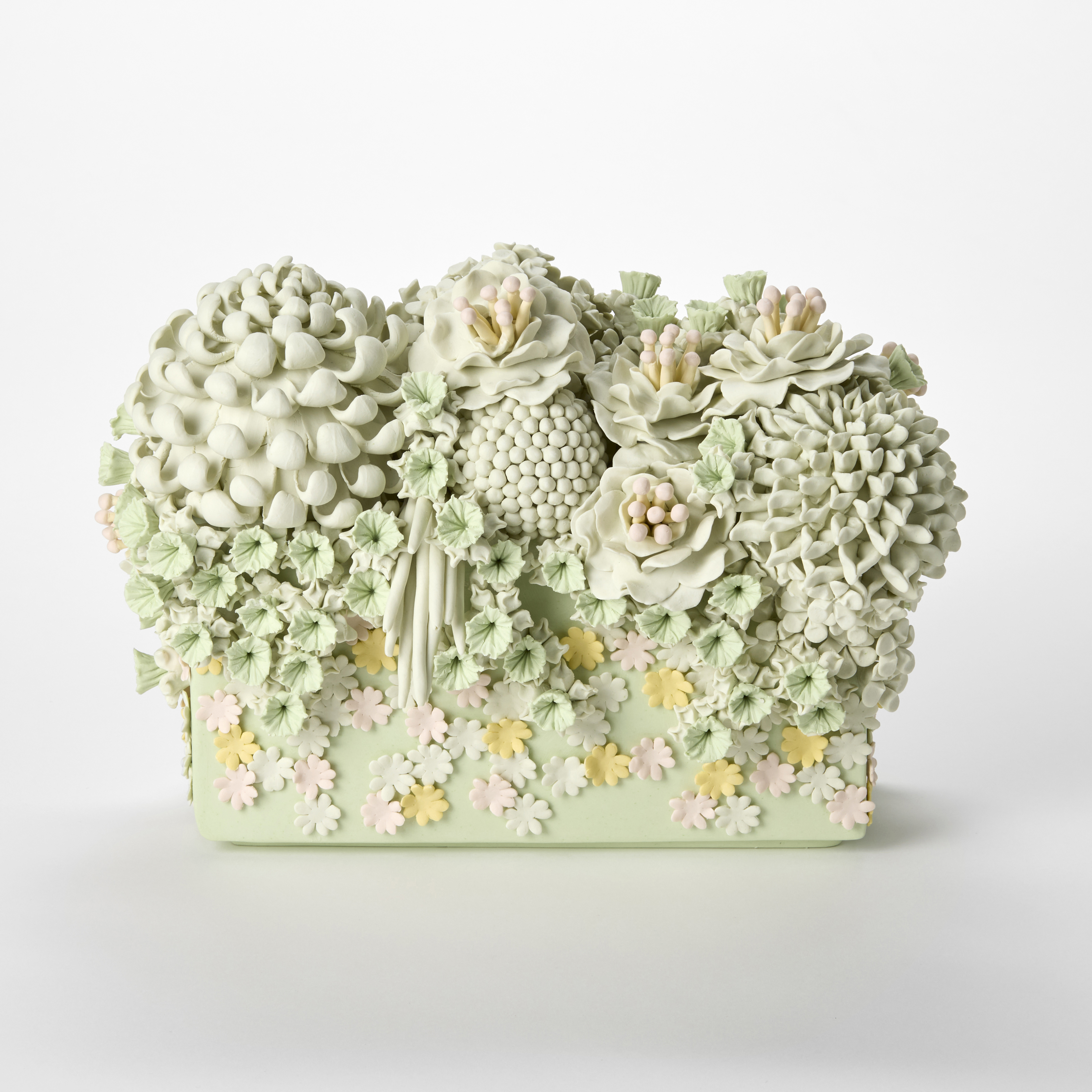 a pastel coloured ceramic sculpture resembling a floral arrangement featuring intricately sculpted flowers and textured blossoms in shades of pale green white and soft pink densely covering the top and front of a rectangular base arranged in a symmetrical decorative pattern evoking the look of piped icing or ornamental cake decoration hand made from coloured porcelain