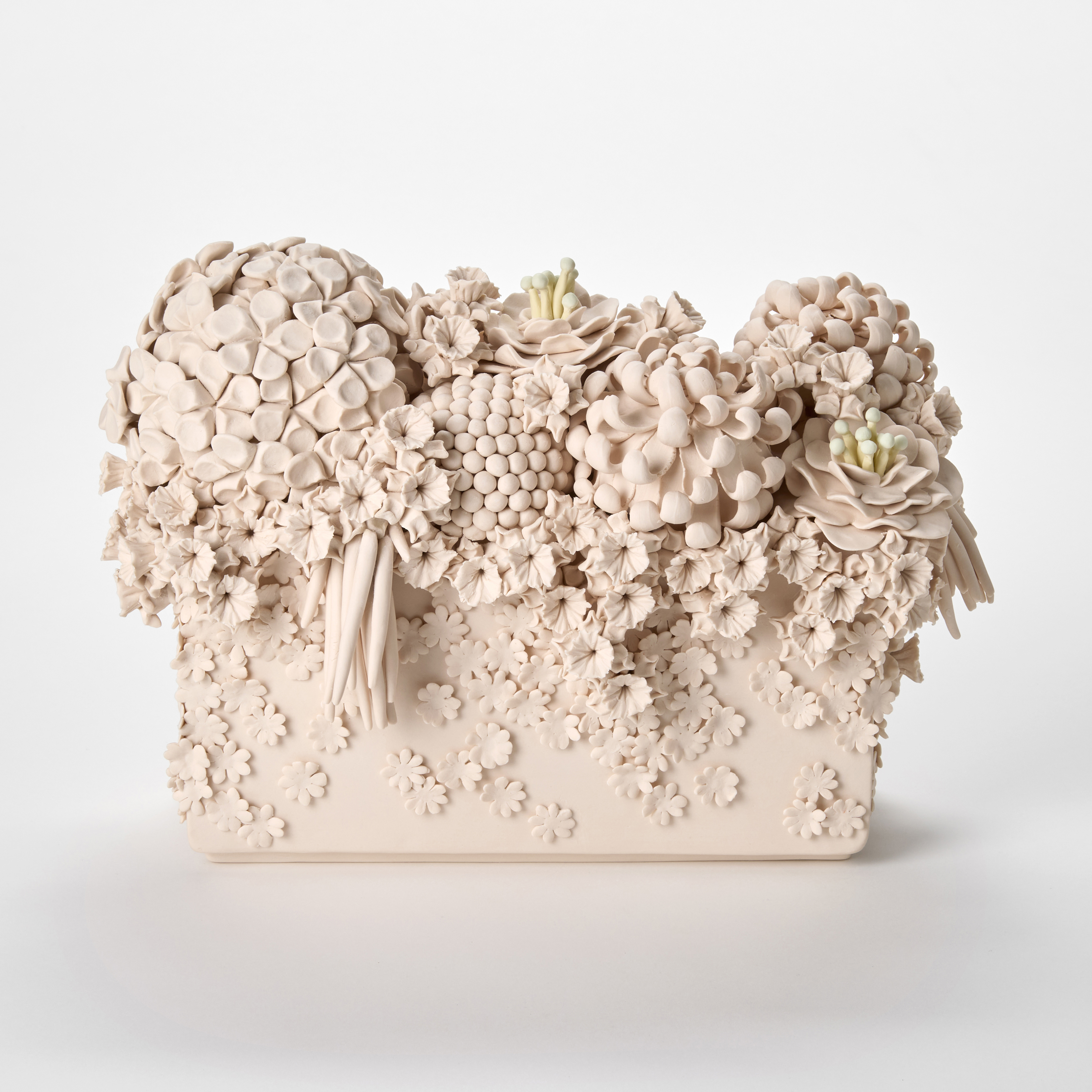 a monochromatic light pink ceramic sculpture resembling a dense floral arrangement featuring an ornate cluster of intricately sculpted flowers including hydrangea like blooms roses and textured spherical elements overflowing from the top of a rectangular base with smaller blossoms cascading down the sides giving the piece a highly detailed cake-like decorative appearance hand made from coloured porcelain