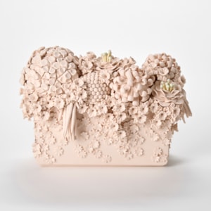 a monochromatic light pink ceramic sculpture resembling a dense floral arrangement featuring an ornate cluster of intricately sculpted flowers including hydrangea like blooms roses and textured spherical elements overflowing from the top of a rectangular base with smaller blossoms cascading down the sides giving the piece a highly detailed cake-like decorative appearance hand made from coloured porcelain