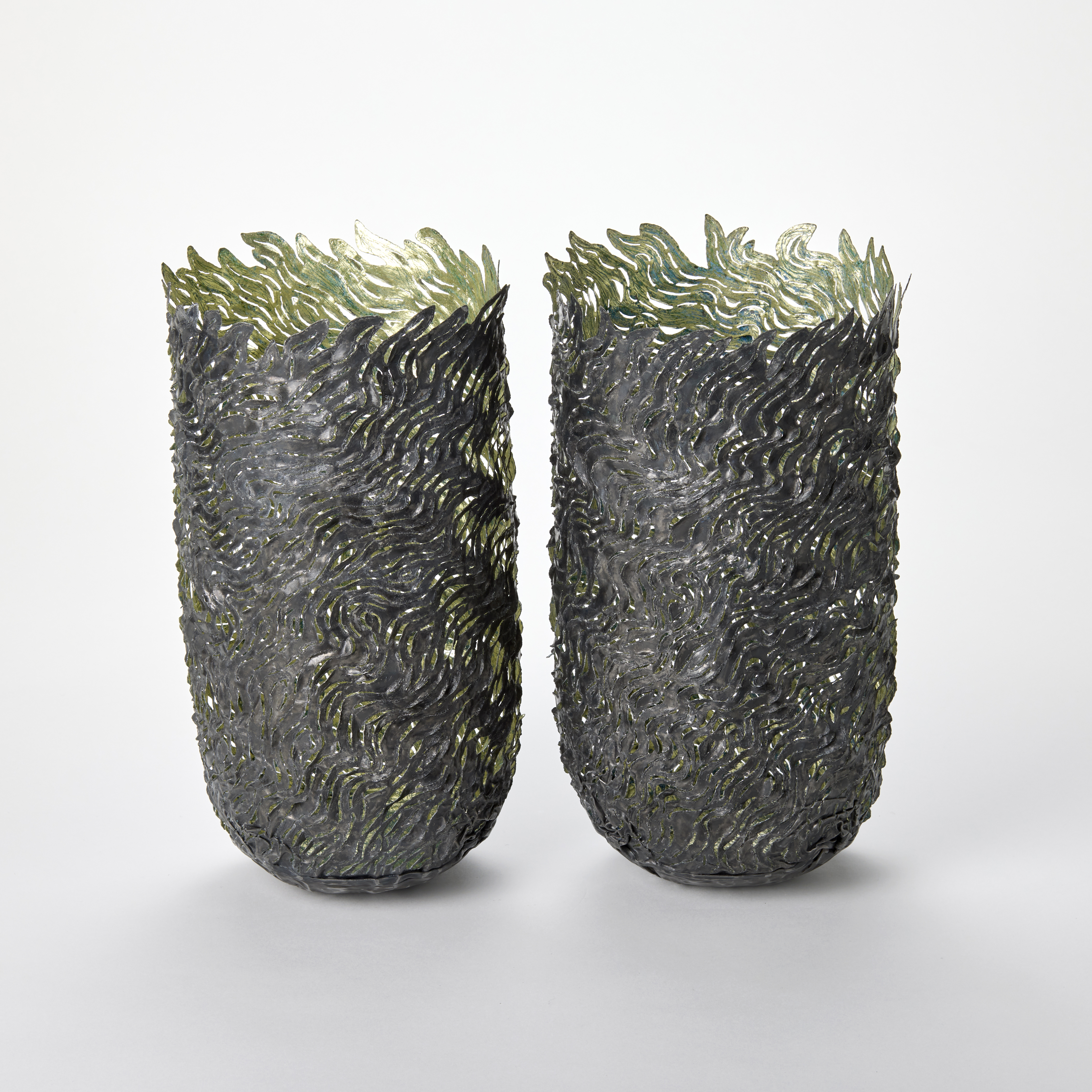 a pair of tall cylindrical patinated steel vessels with textured wave-like patterns covering the surface with each predominantly in dark gray with subtle hints of green and metallic sheen with irregular tops and decorated with flame-like leafy protrusions in green tones giving the pieces an organic almost underwater or botanical appearance hand made from steel and gold leaf