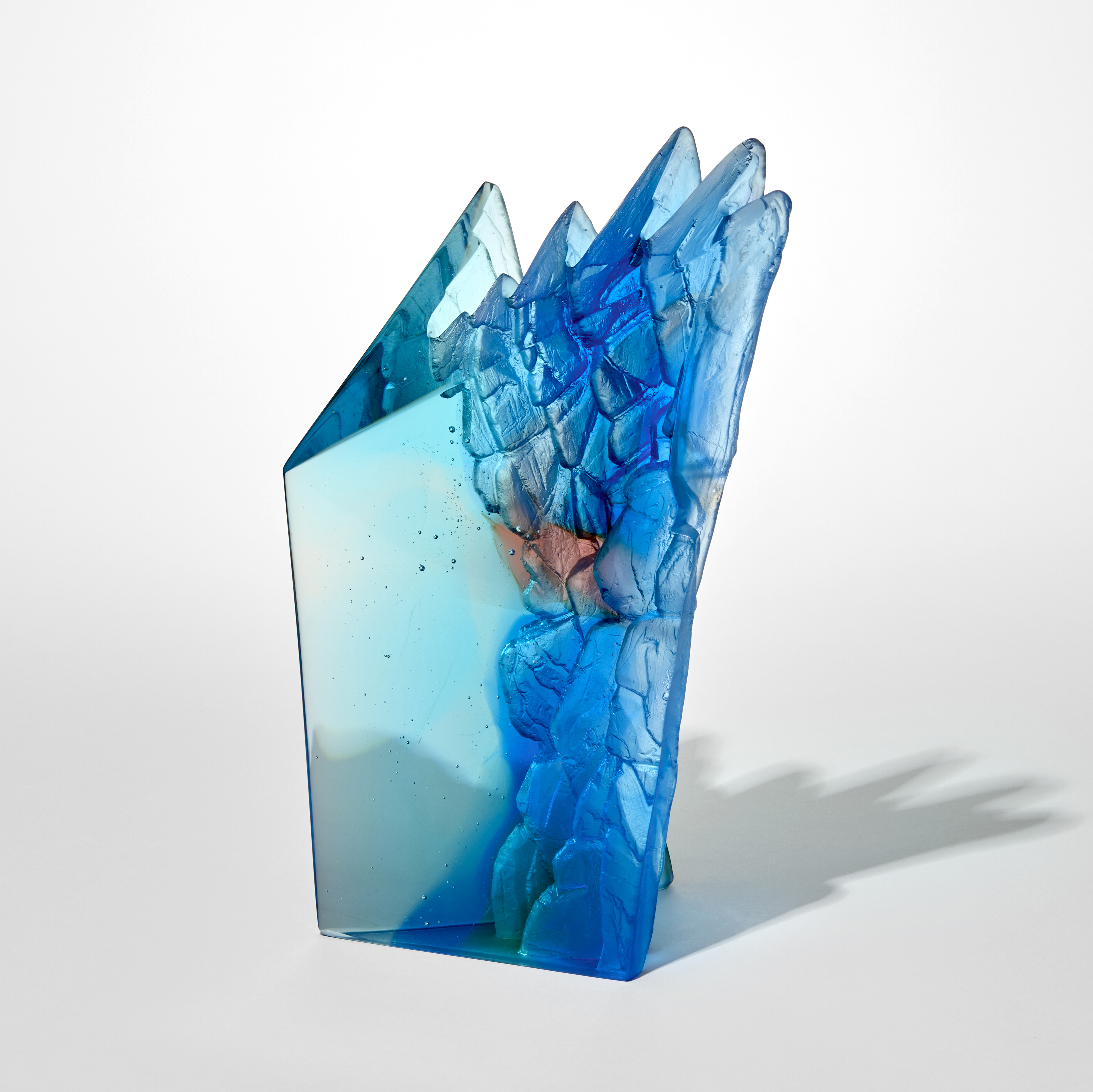 tall angular transparent abstract jade blue and pink sculpture with two flat and polished sides and two inverted sides with craggy rock like textures hand made from cast glass  
