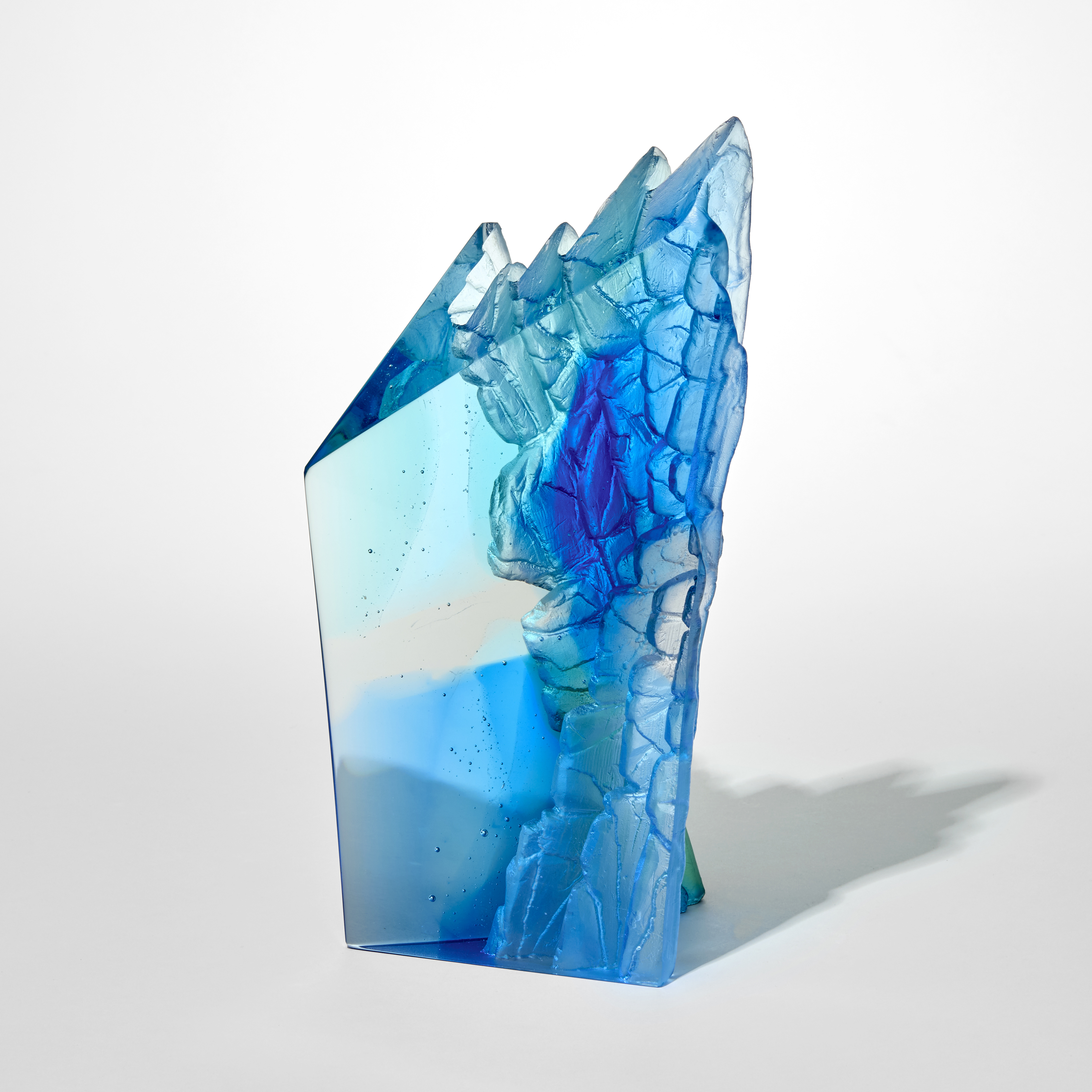 abstract transparent sculpture in shades of blue shaped like a tall angular block with a jagged crystalline top edge and with one smooth and polished side and the one textured and craggy resembling rock formations hand made from cast glass