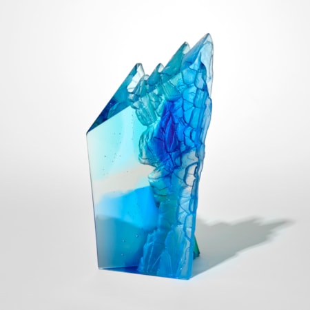 abstract transparent sculpture in shades of blue shaped like a tall angular block with a jagged crystalline top edge and with one smooth and polished side and the one textured and craggy resembling rock formations hand made from cast glass