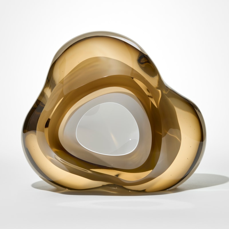 smooth abstract geode inspired sculpture with an organic rounded triangular form created from layers of translucent clear amber and brown glass with a central opaque white core surrounding a central hollow hand made from blown and sculpted glass