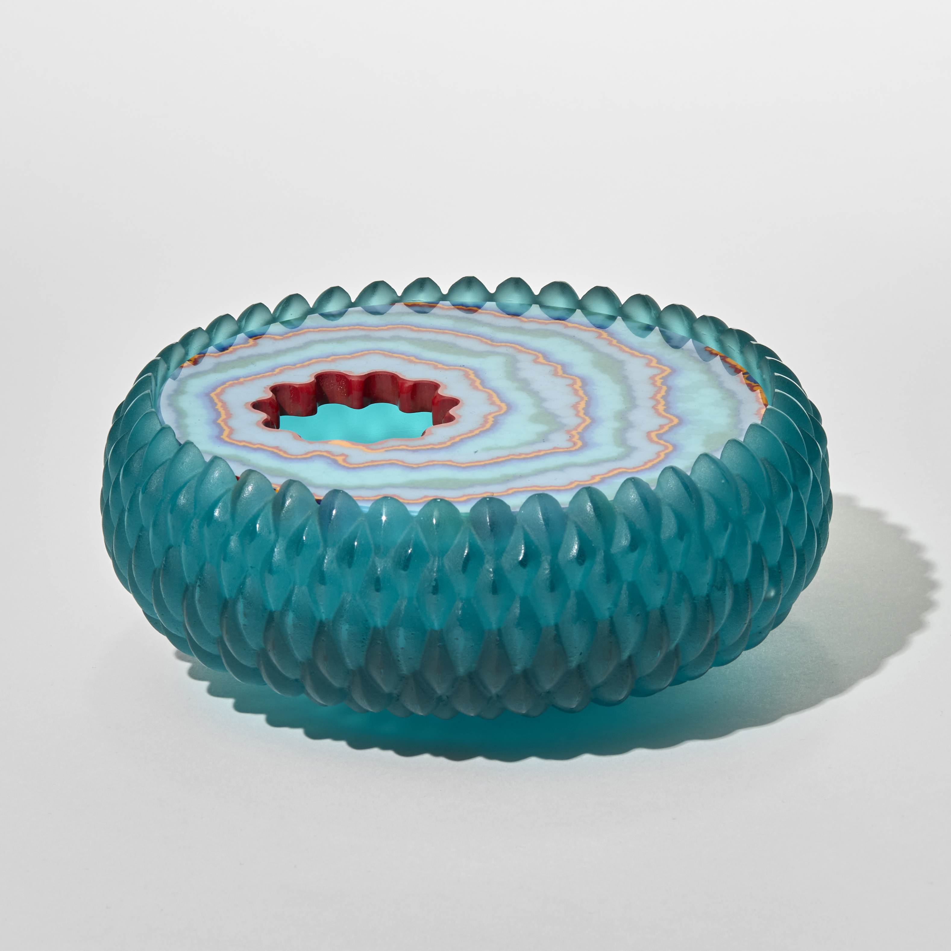 a circular low glass lidded box with a textured raised cross hatched teal exterior resembling scales or droplets and a lid featuring concentric patterned rings in shades of blue white and red surrounding a round off-set opening with an undulating edge with the appearance of a gemstone and its translucent qualities hand made from cast glass