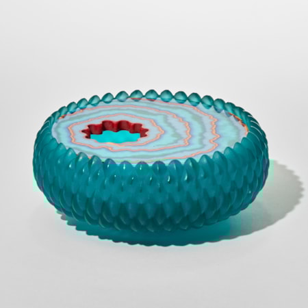 a circular low glass lidded box with a textured raised cross hatched teal exterior resembling scales or droplets and a lid featuring concentric patterned rings in shades of blue white and red surrounding a round off-set opening with an undulating edge with the appearance of a gemstone and its translucent qualities hand made from cast glass