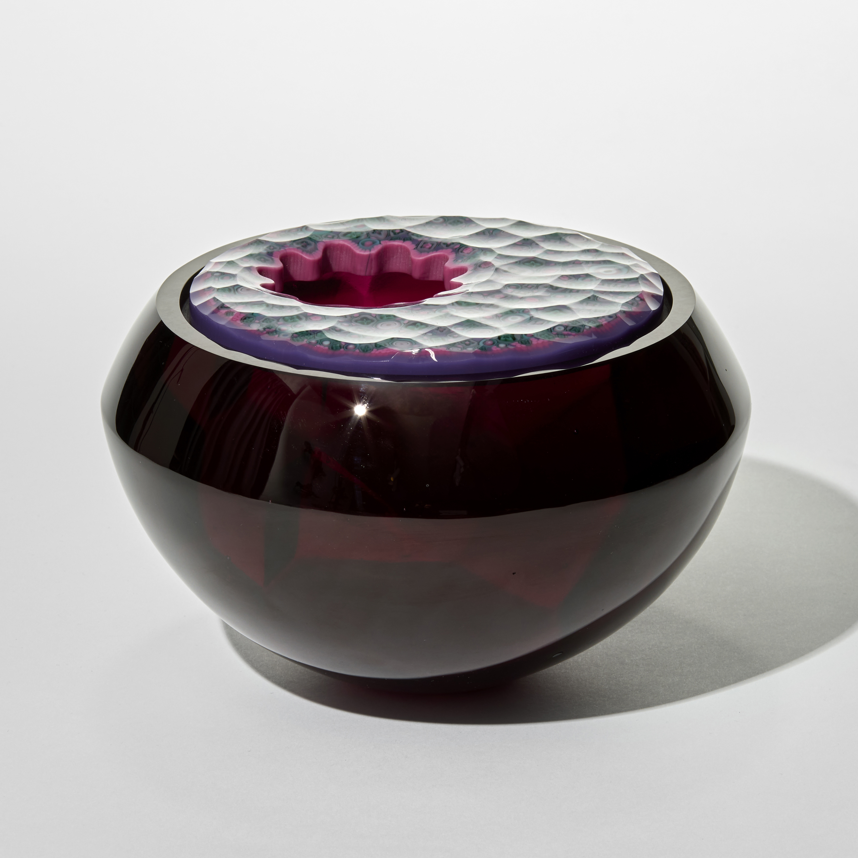 rounded glass facetted box in deep opaque plum with a smooth reflective surface and an inserted lid with with a faceted top surface incorporating repeat tiny intricate patterns in pink purple and jade with an off centre opening with a scalloped edge hand made from cast glass