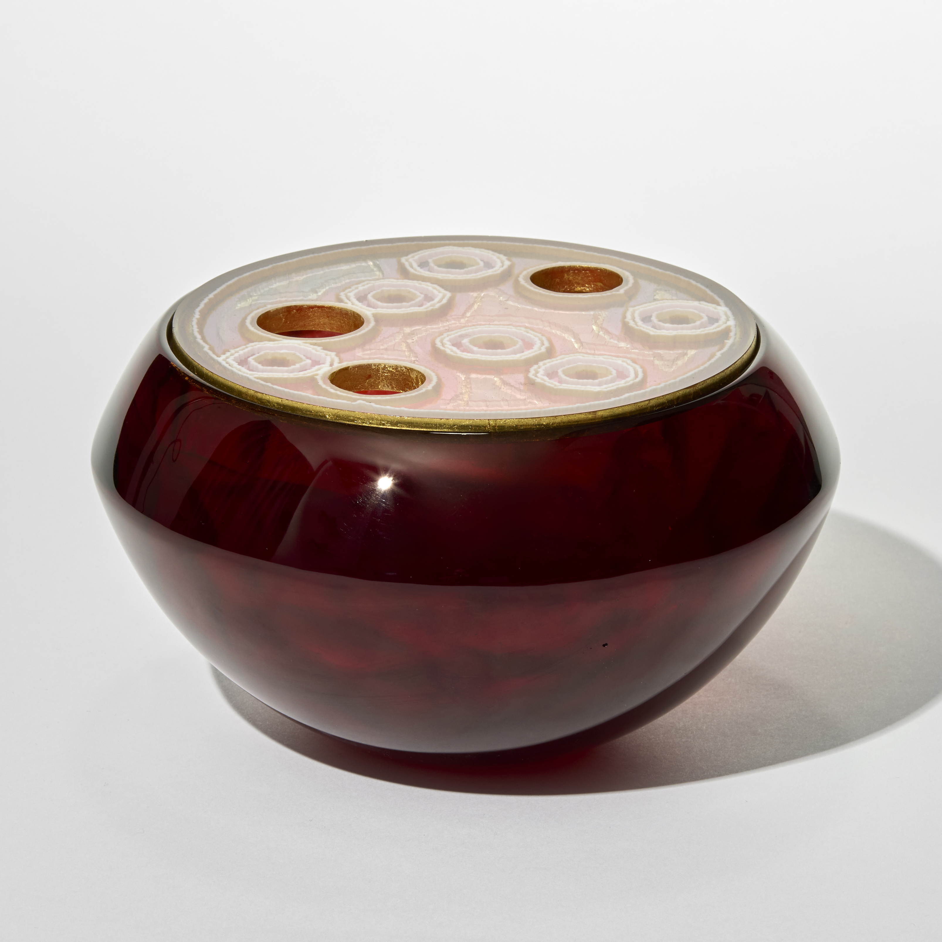 facetted round glass box in deep opaque toffee brown with a smooth polished outer surface and a lid with pale pink amber and gold concentric circular motifs with some solid with frilly white dotted rings and some cutouts in varying sizes creating a patterned lotus seed like effect hand made from cast glass