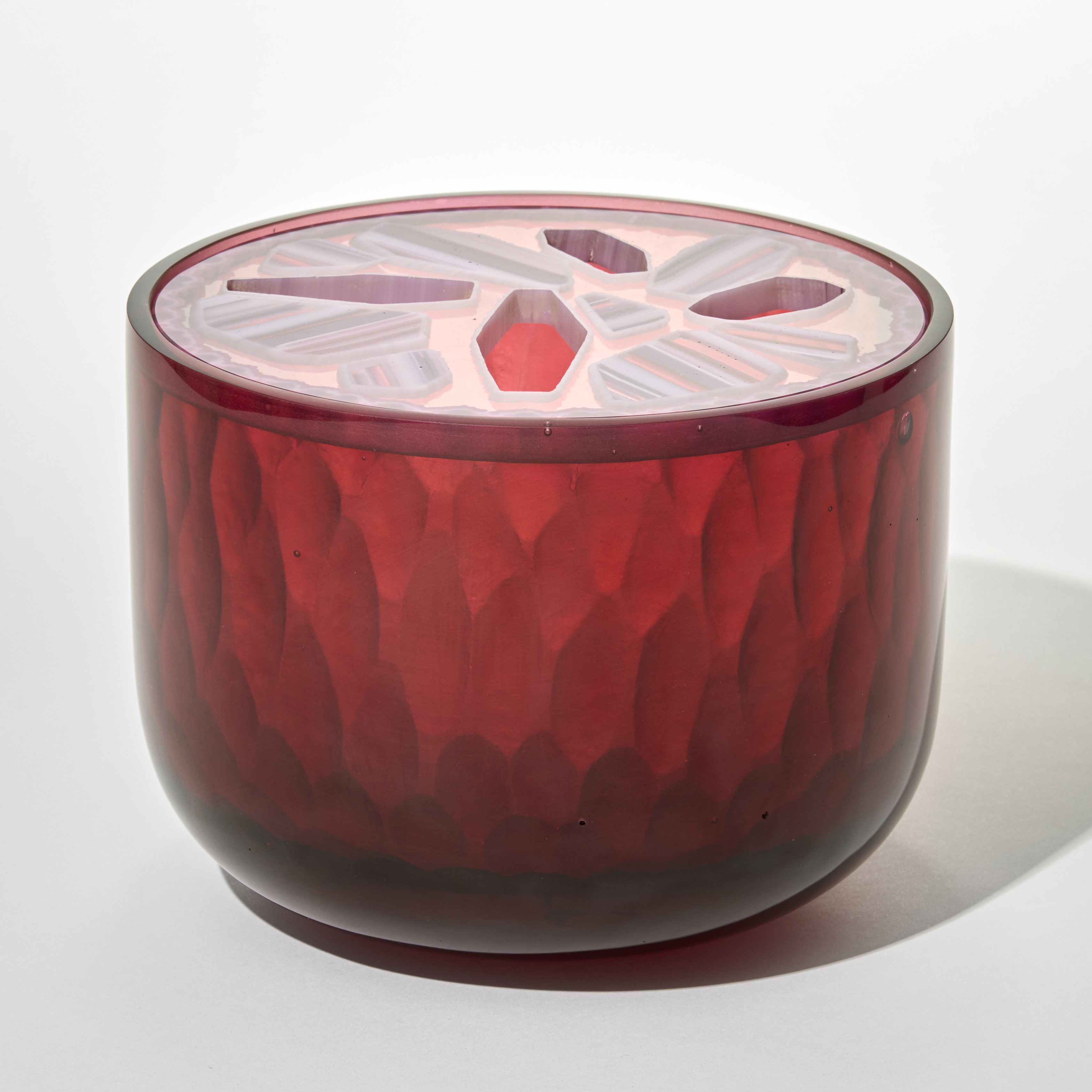 cylindrical glass opaque box in deep pinky red with a faceted semitransparent lid that is patterned with pale pink and white geometric gem shapes with some solid and some cutouts forming star like shapes hand made from cast glass