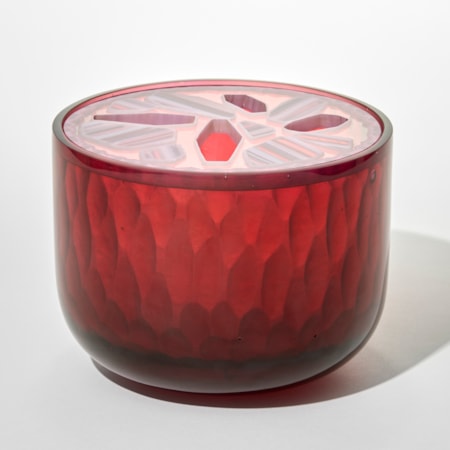 cylindrical glass opaque box in deep pinky red with a faceted semitransparent lid that is patterned with pale pink and white geometric gem shapes with some solid and some cutouts forming star like shapes hand made from cast glass