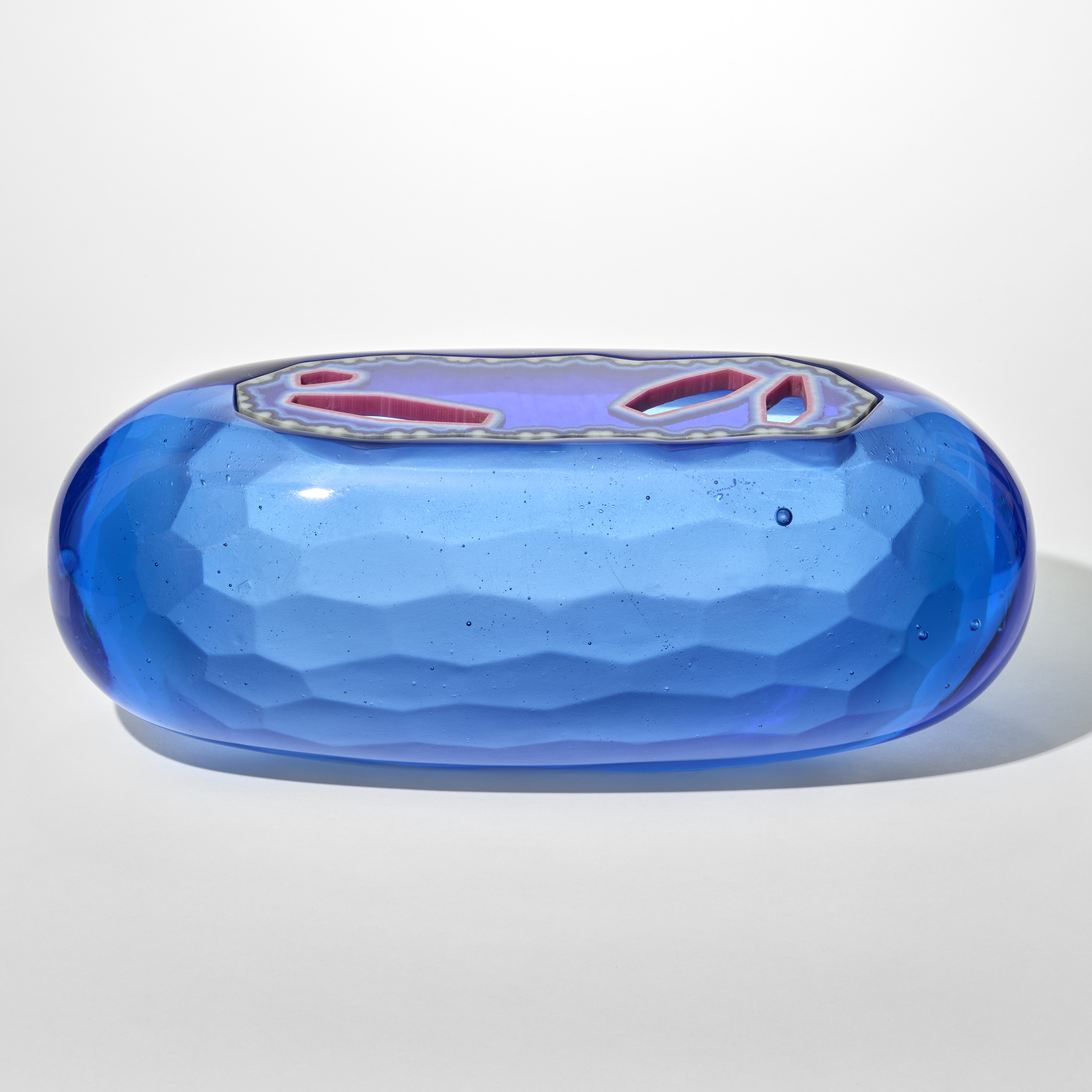 a smooth capsule shaped opaque glass sculpture in vibrant blue with a faceted inserted lid that features irregular red and clear geometric openings giving the piece a jewel like translucent quality with a textured tessellating interior surface hand made from cast glass