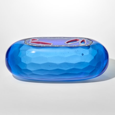 a smooth capsule shaped opaque glass sculpture in vibrant blue with a faceted inserted lid that features irregular red and clear geometric openings giving the piece a jewel like translucent quality with a textured tessellating interior surface hand made from cast glass