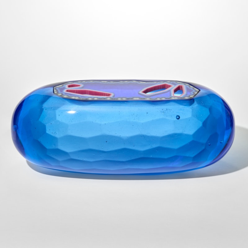 a smooth capsule shaped opaque glass sculpture in vibrant blue with a faceted inserted lid that features irregular red and clear geometric openings giving the piece a jewel like translucent quality with a textured tessellating interior surface hand made from cast glass