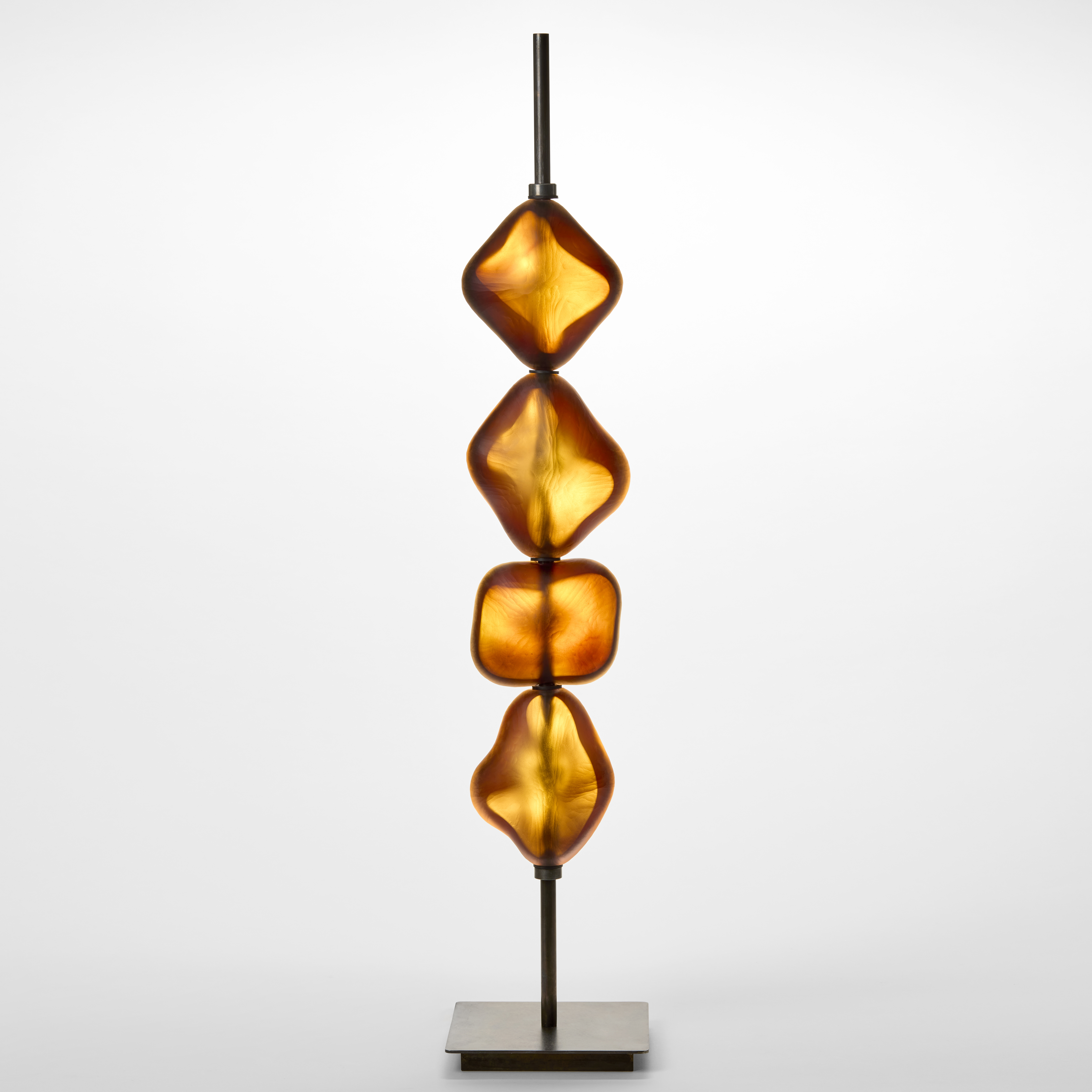 tall metal pole with a square base and four geometric opaque shapes in amber suspended on the stem with each containing an LED light directing the light behind it hand blown from glass with a textured cut surface