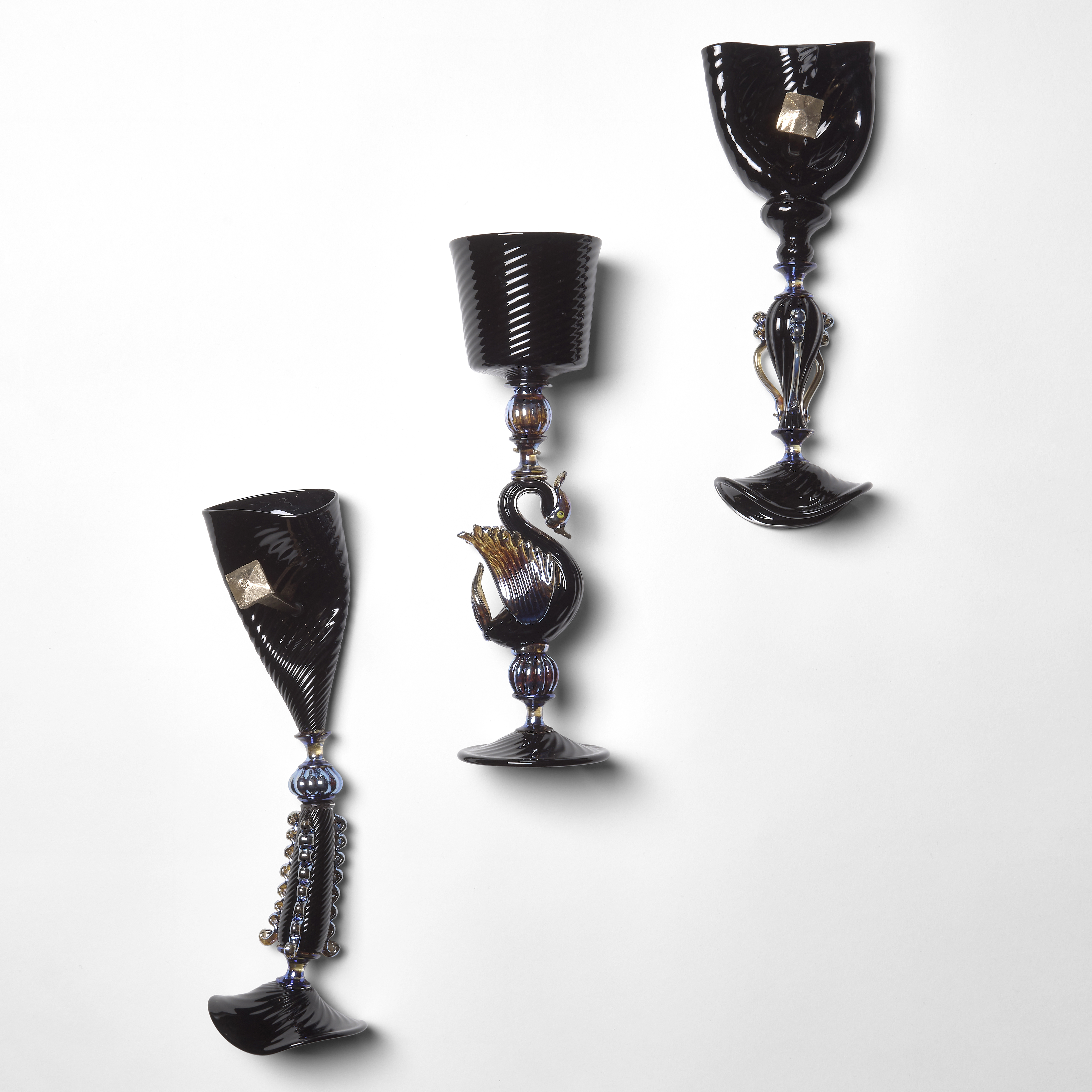 three glossy black goblets suspended on a wall with the inner one perfect held by a refined fitting and the outer two squashed and damaged held in place by large nails hand sculpted from glass