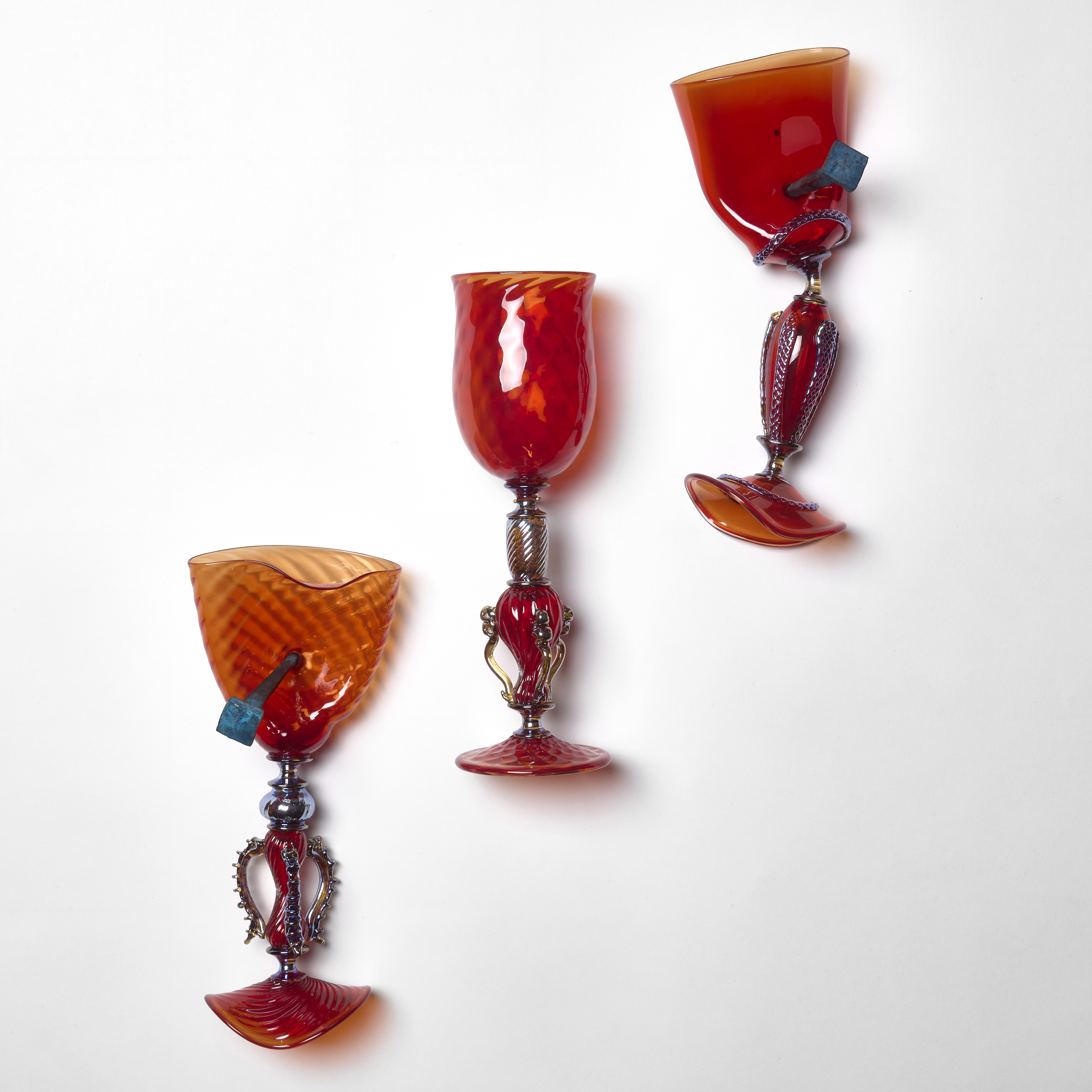 diagonally hanging wall installation consisting of three bright scarlet red venetian inspired detailed goblets with the middle one perfect and on an elegant fitting with the outer two squashed and distorted nailed in place by two chunky bronze nails hand made from sculpted glass
