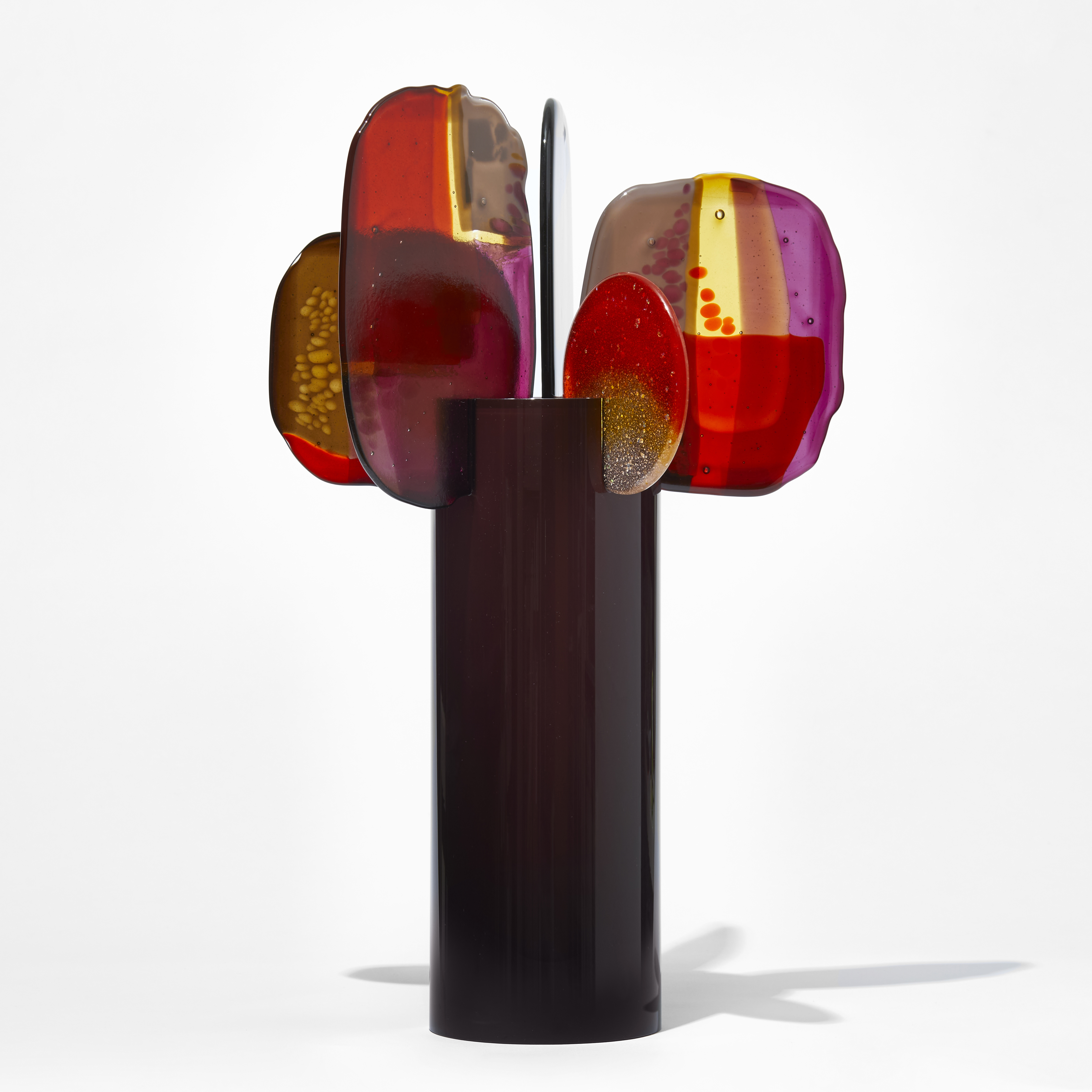 glossy dark chocolate cylinder with five rounded finials of glass perched on the top edge each with abstract patterns in bright red soft yellow purple and deep pink hand made from blown and fused glass