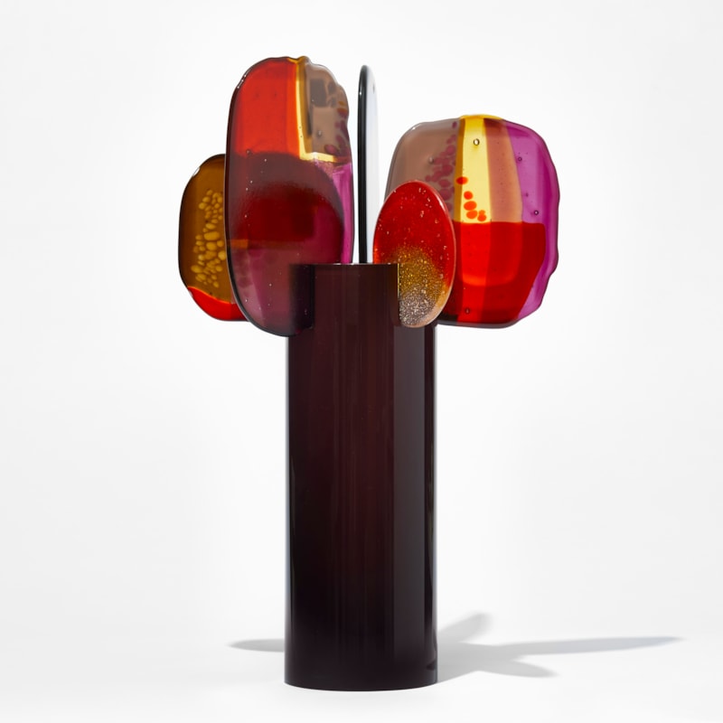 glossy dark chocolate cylinder with five rounded finials of glass perched on the top edge each with abstract patterns in bright red soft yellow purple and deep pink hand made from blown and fused glass