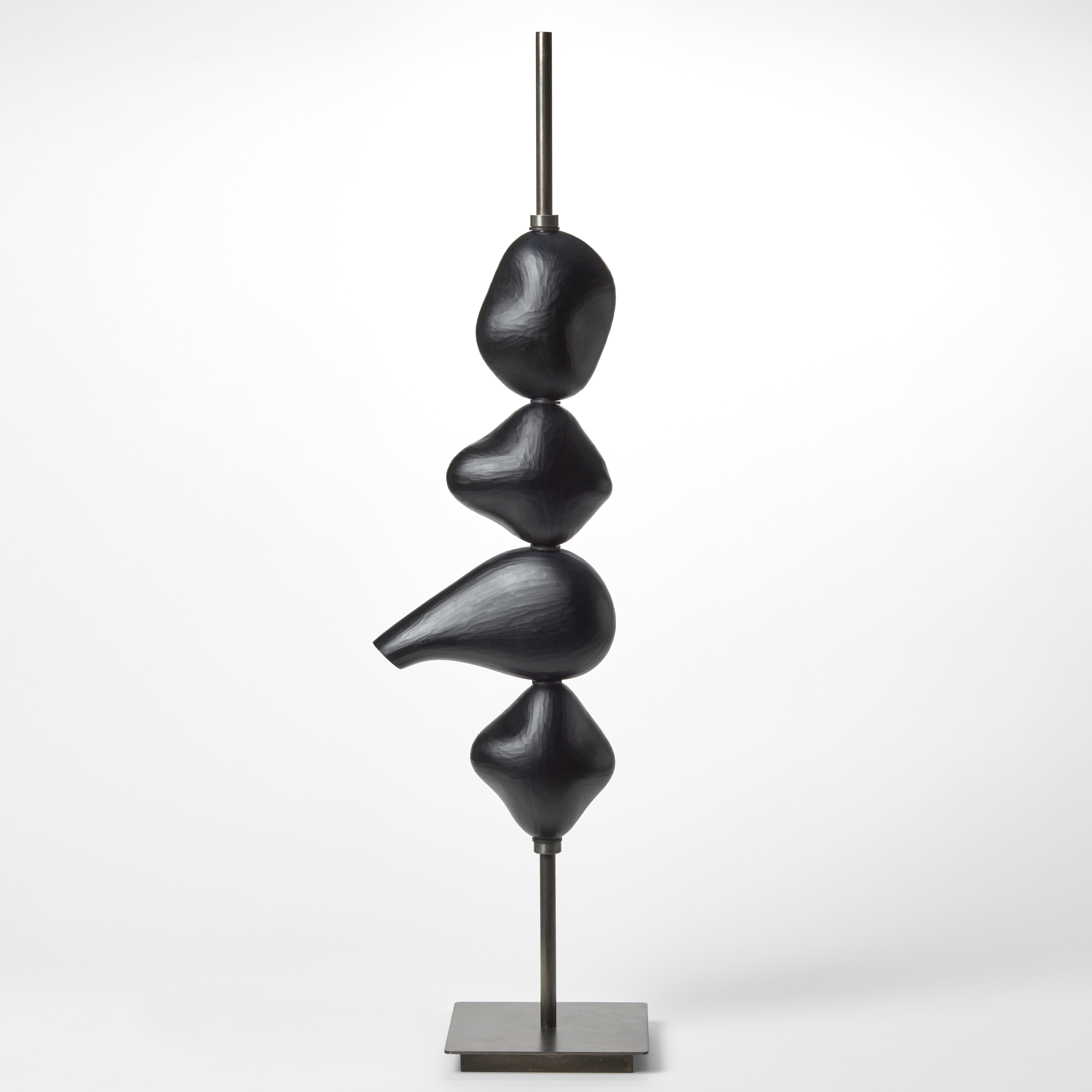sculptural light with a steel square base and long thin pole which suspends four black abstract shapes with each containing an LED light hand made from blown glass with a repeat textured surface