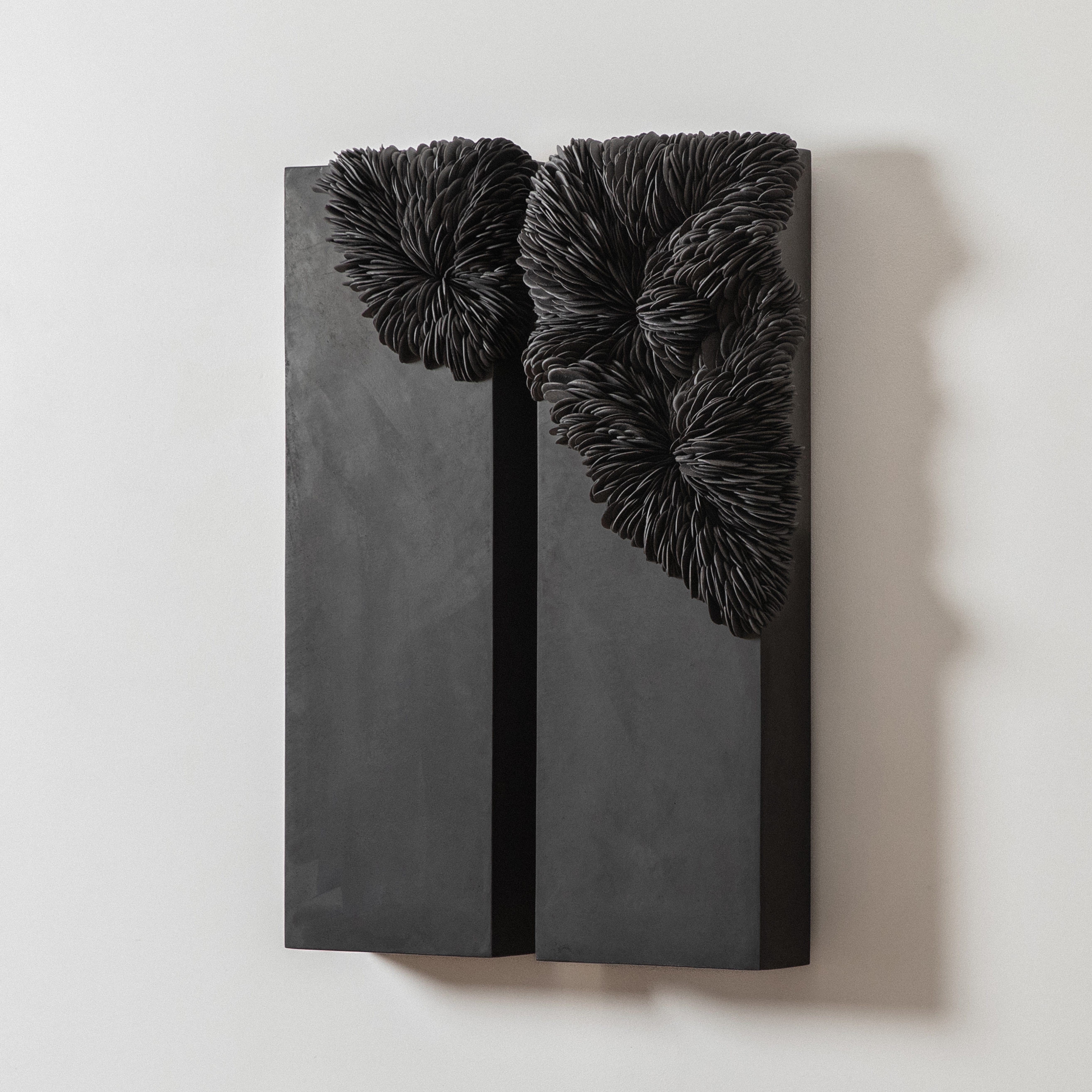 two long substantial rectangular chunky black panels that are hung on the wall with the upper triangular third embedded with curved interlocking shards traversing from one panel to the other made from porcelain and tadelakt plaster