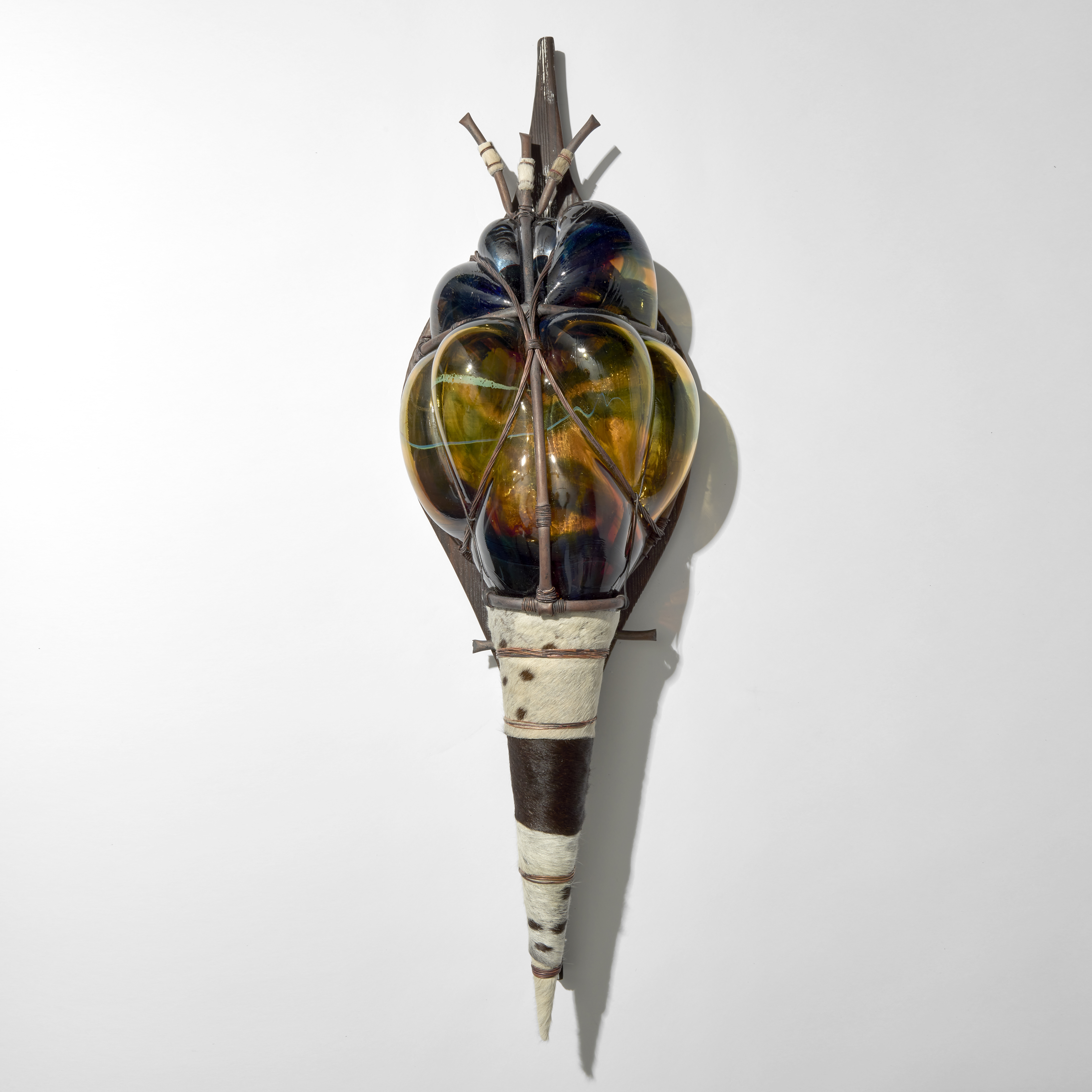 stylised african wall mounted shield created from a copper cage with merged green and blue blown glass bulging through the gaps and a tapering pointed base wrapped in cow hide with the entire object mounted on a sympathetically shaped wooden board